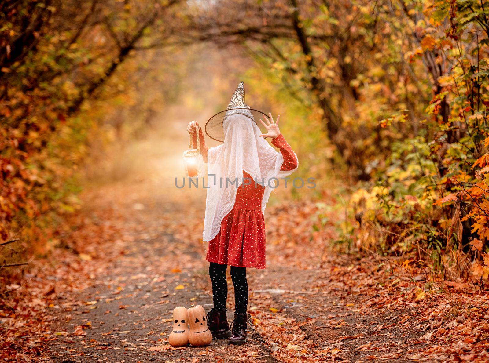 Little girl dressed for halloween party standing in autumn wood near decorated pumpkins and flying bats