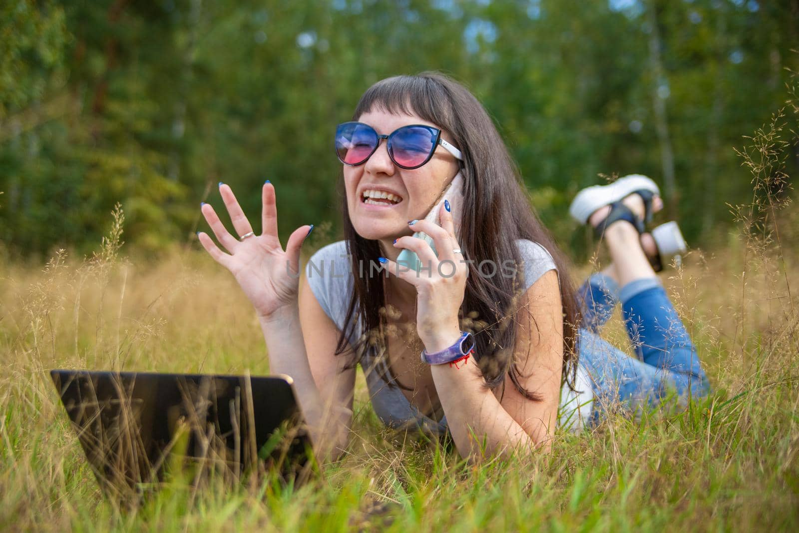 young woman swears on the phone while looking into a laptop. woman freelancer works in a sunny meadow