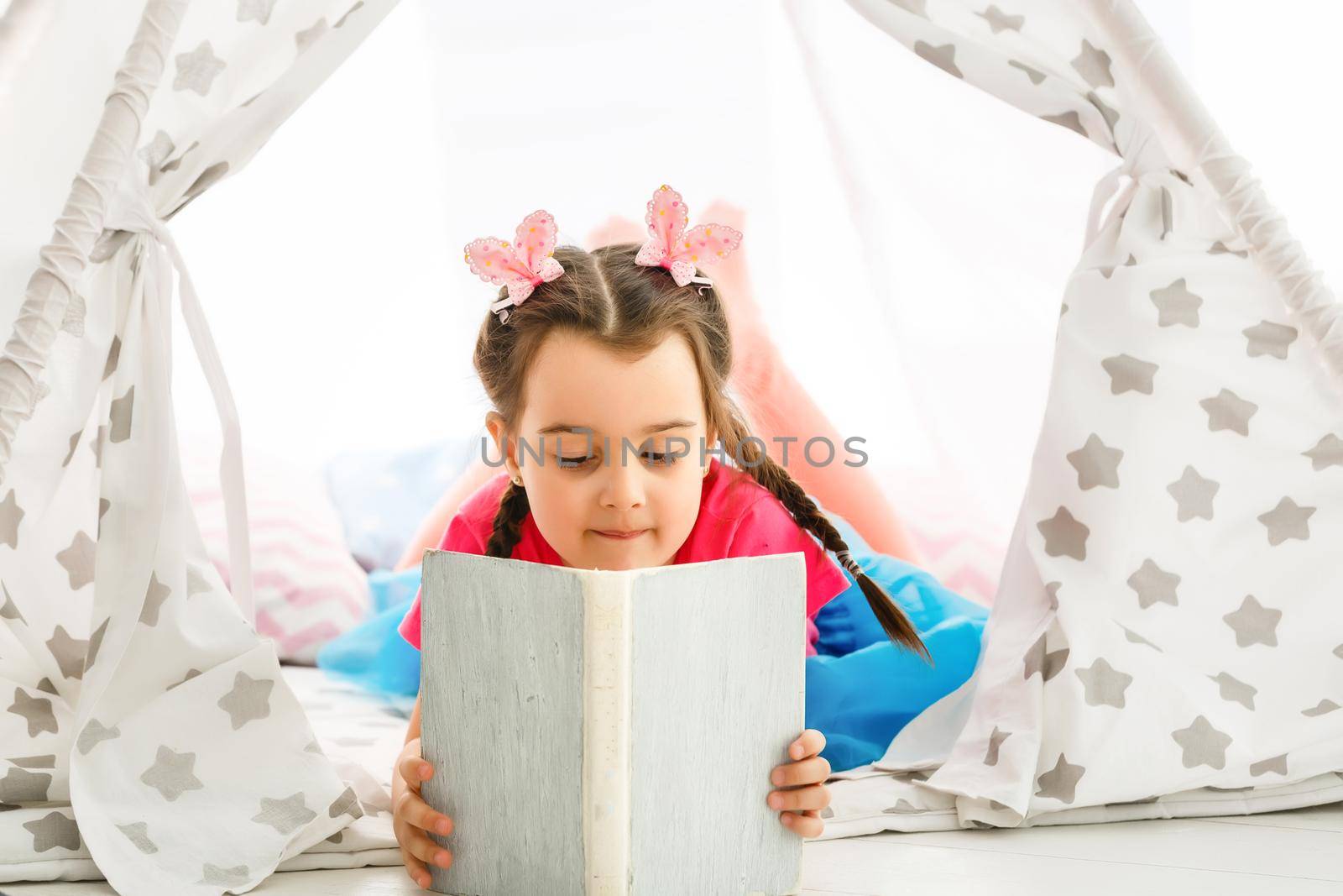 Enjoying great story. Cute little girl reading book while sitting on the floor in tent by Andelov13