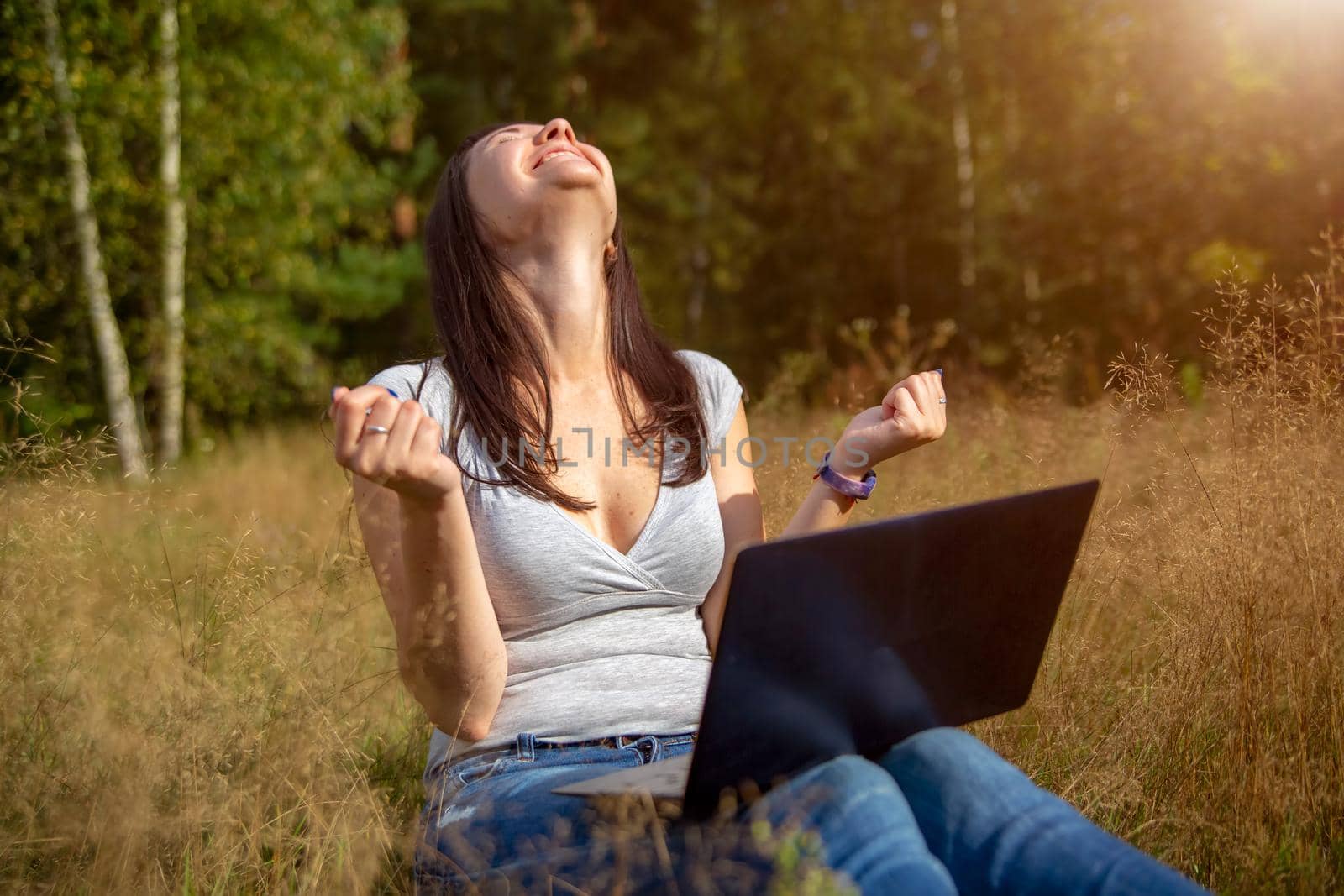 happy young woman with laptop on a sunny lawn. freelancer works in nature. student studies remotely by Mariaprovector
