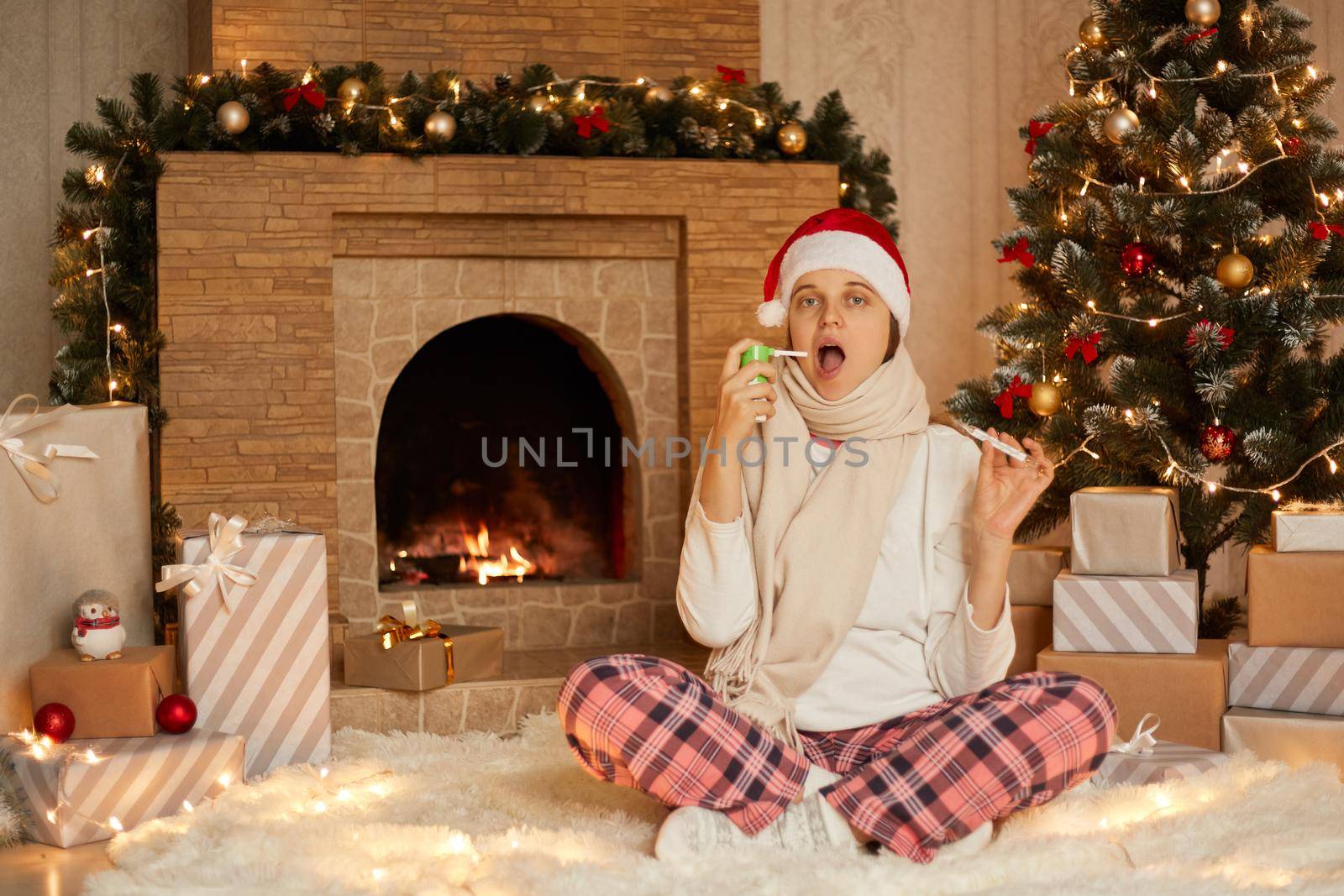 Woman having problem with health, being at home, lady sitting in floor with crossed legs, showing thermometer to camera, using sore throat spray, wearing home closing and santa hat.