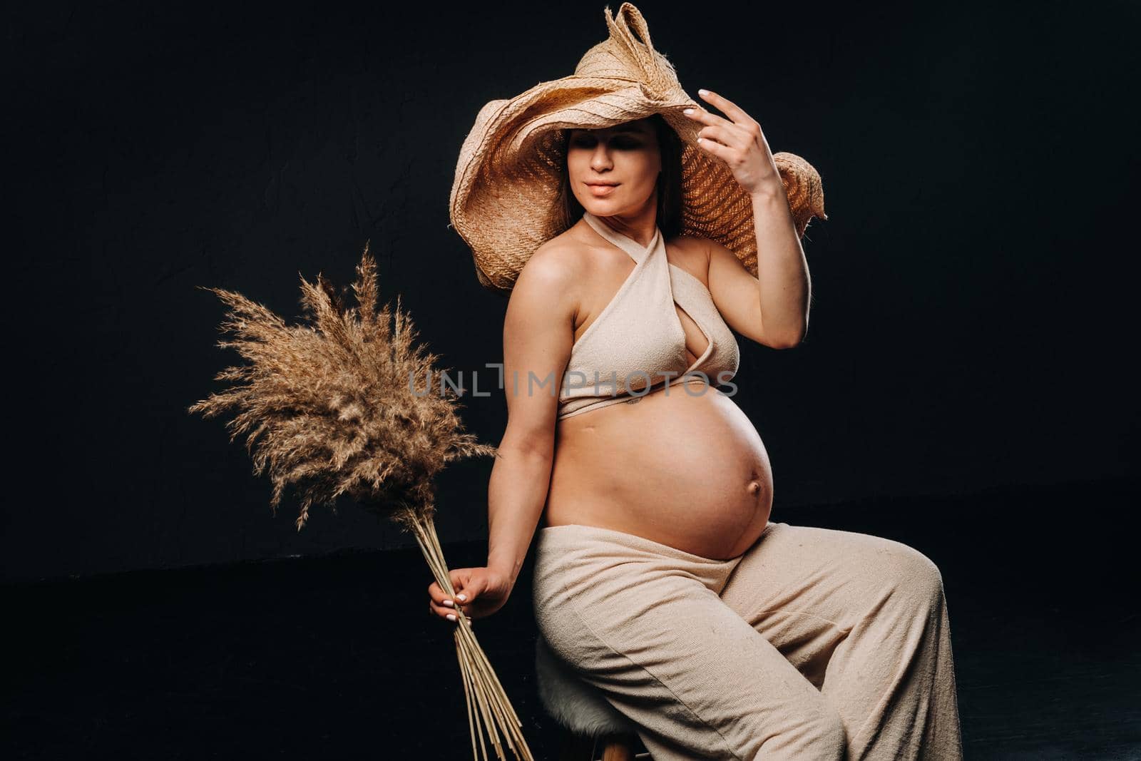 pregnant woman in a straw hat with beige clothes with a bouquet in her hands in the studio on a black background.