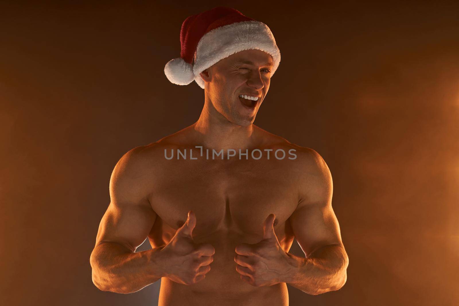 Portrait of muscular man wearing Christmas Santa hat, showing thumb up and smile on smoky background Macho Shirtless Naked torso stripper Hot sexy Santa guy Seductive male for holiday