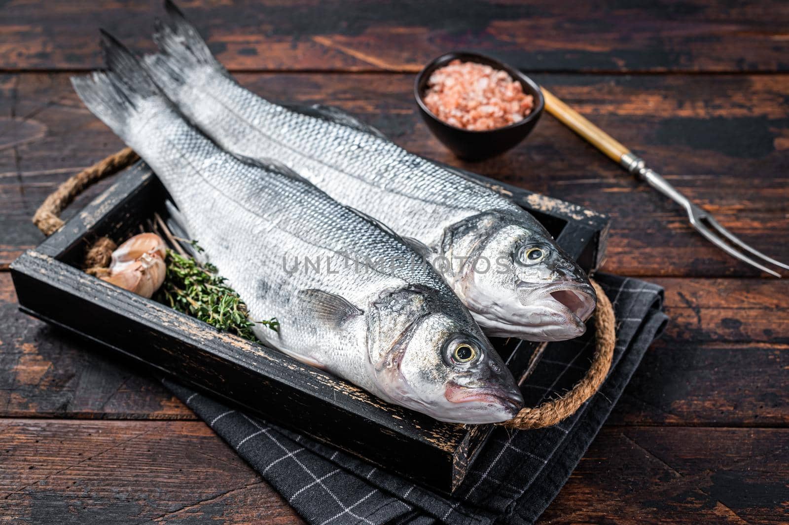 Seabass or sea bass raw fish in a wooden tray with herbs. Dark wooden background. Top view.