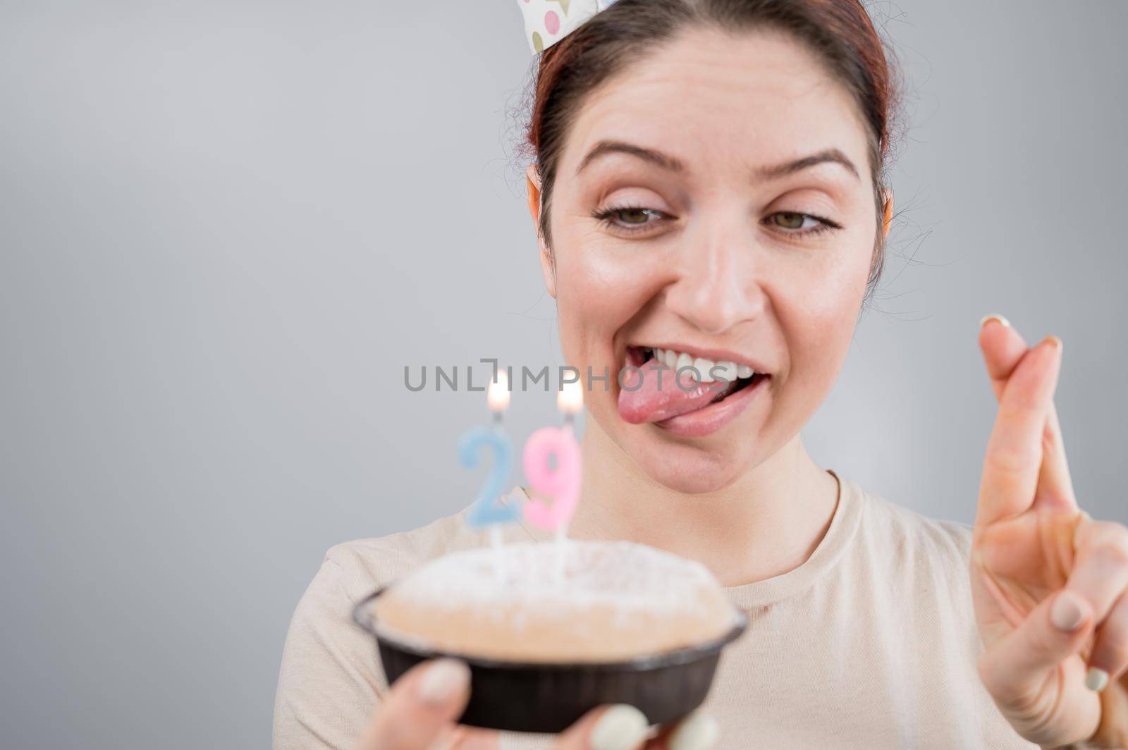 Happy caucasian woman sticking out her tongue and blowing out the candles on the cake with her fingers crossed. The girl celebrates her 29th birthday.