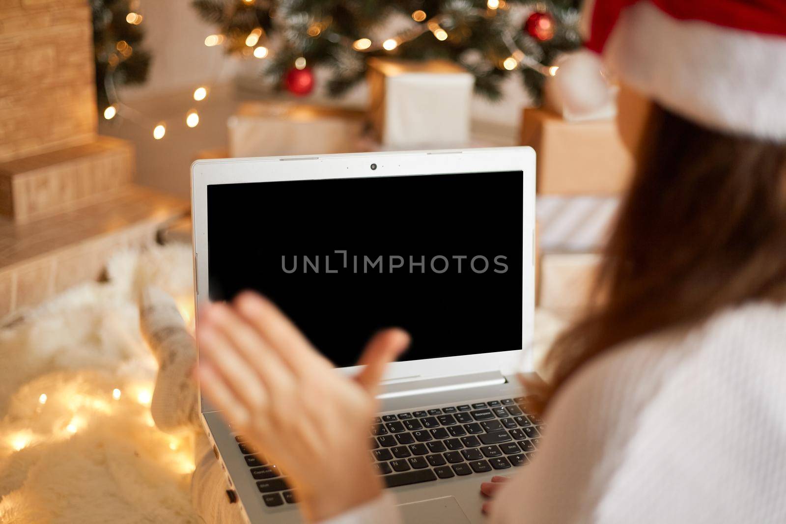 Photo of charming lady sitting and holding laptop, waving palm to screen, wearing santa hat and white sweater in decorated x-mas living room, backwards view of female having video call.