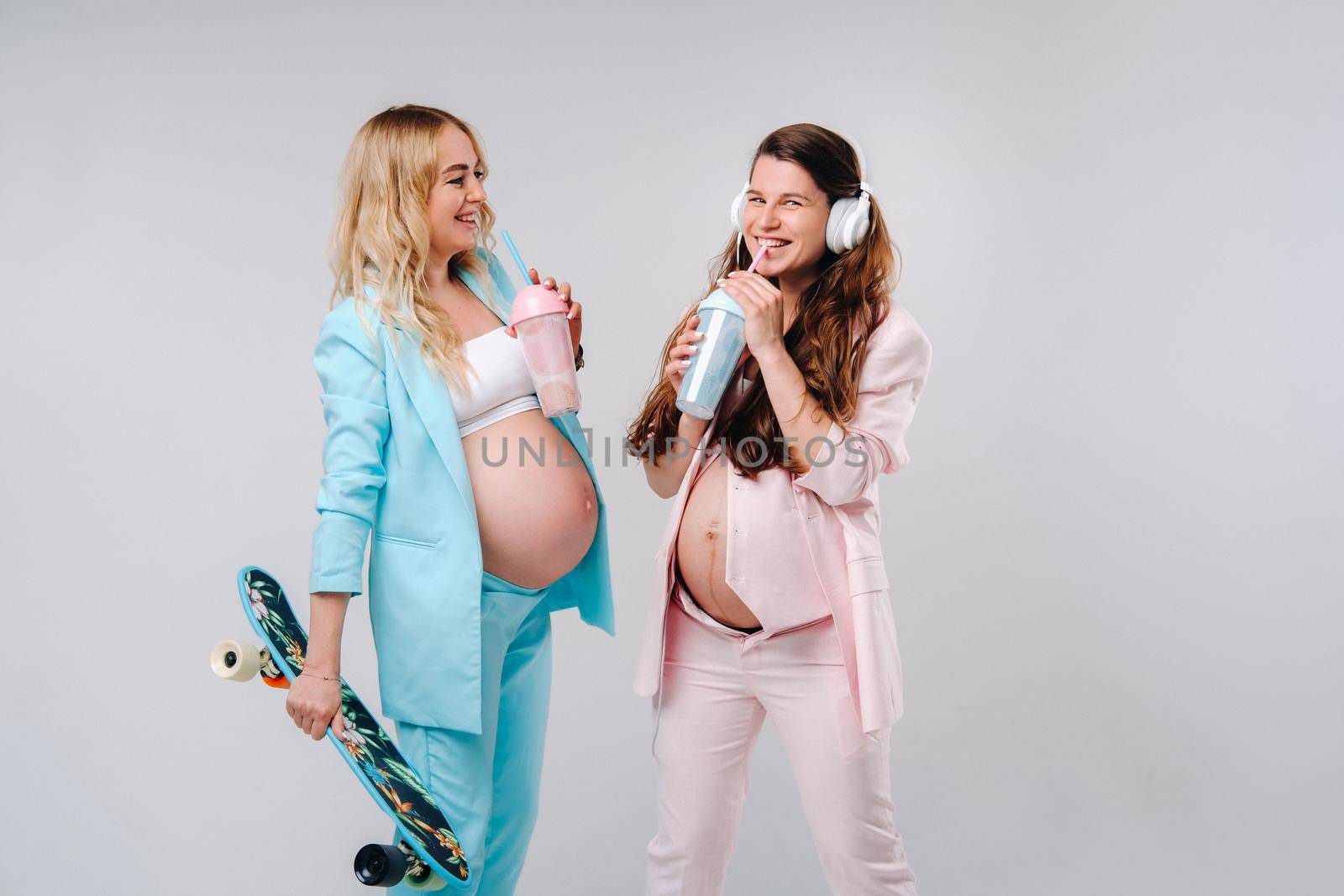 Two pregnant girls in turquoise and pink suits with glasses of juice, a skate and headphones stand on a gray background.