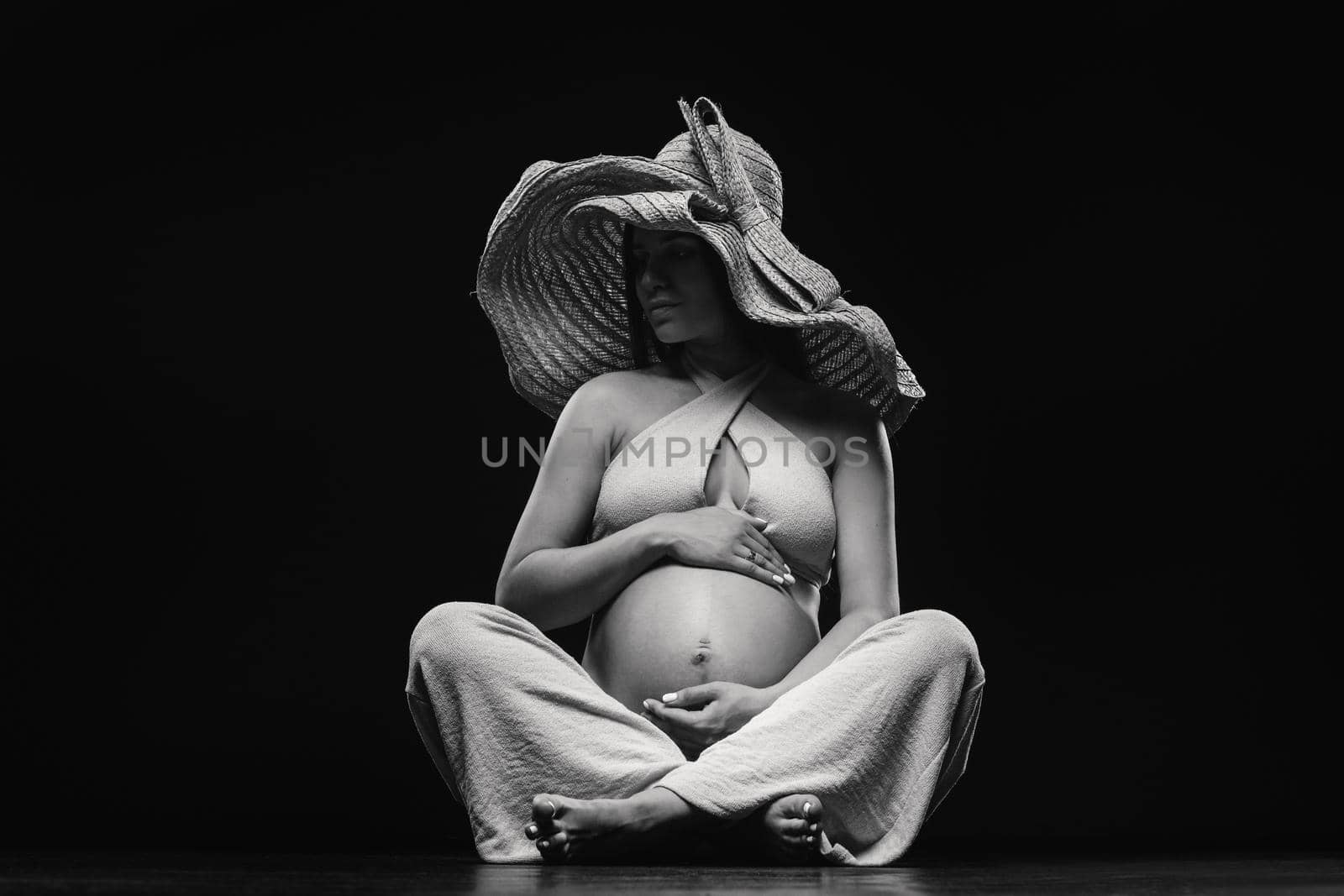 a pregnant woman in a straw hat sits on the floor in beige clothes in a studio on a black background.