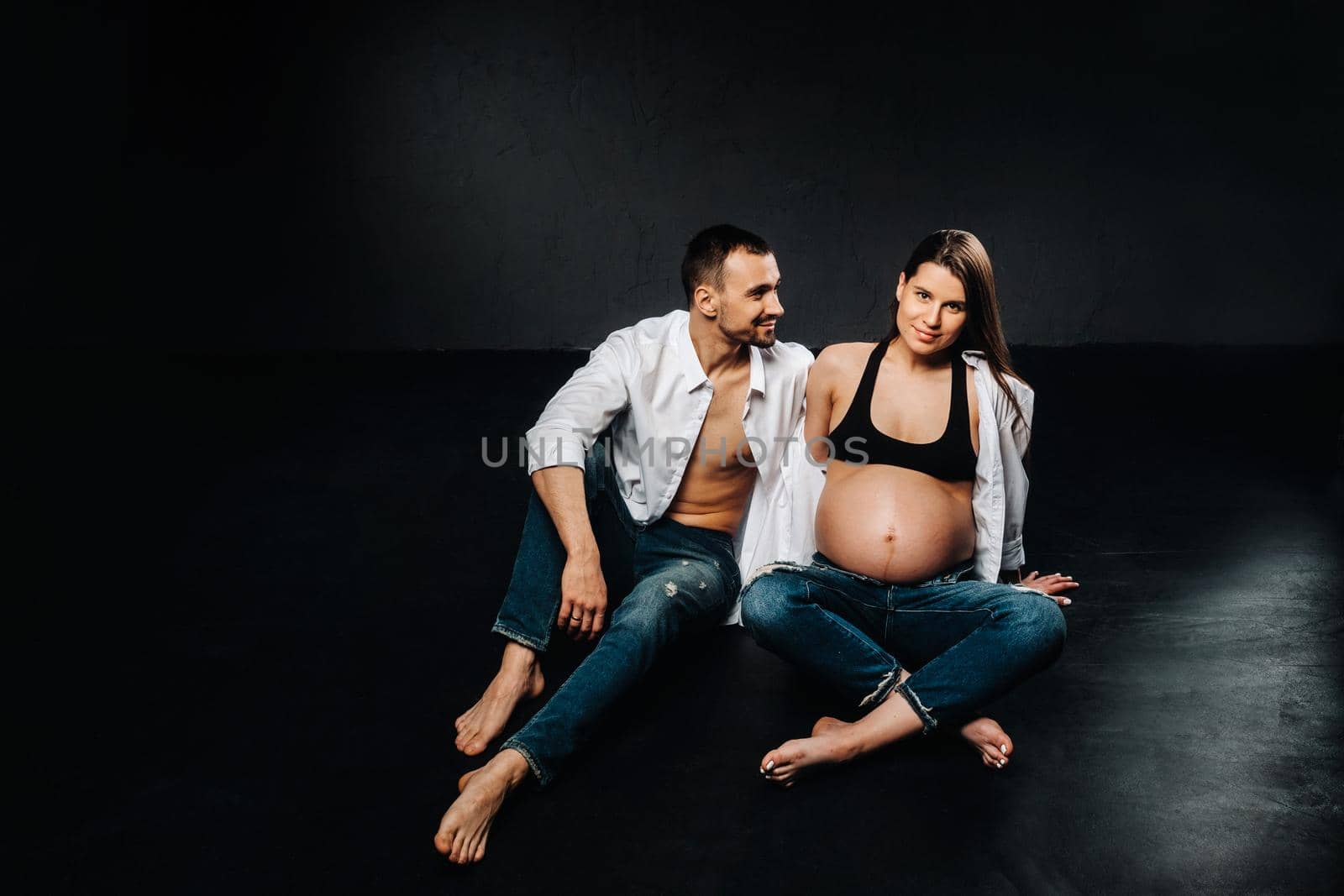 a pregnant woman and a man in a white shirt and jeans in a studio on a black background.