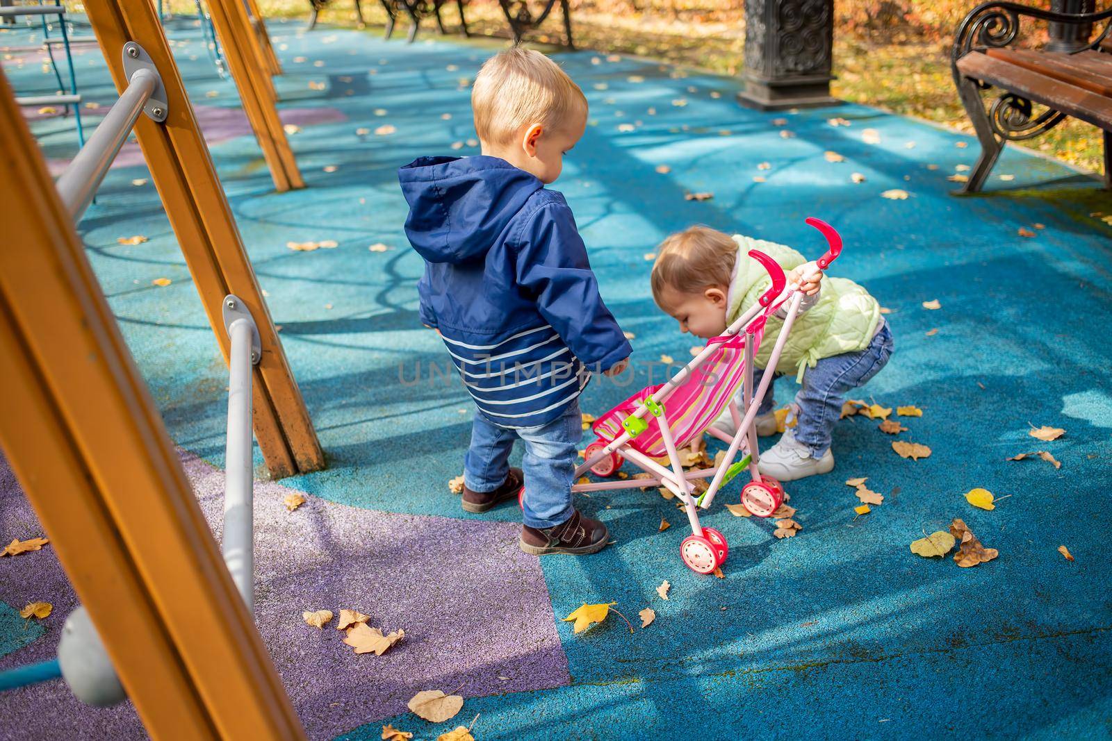 little cute toddler boy and a beautiful year-old baby girl in blue clothes play in a pink toy stroller for a doll in the autumn park in the playground on a sunny day
