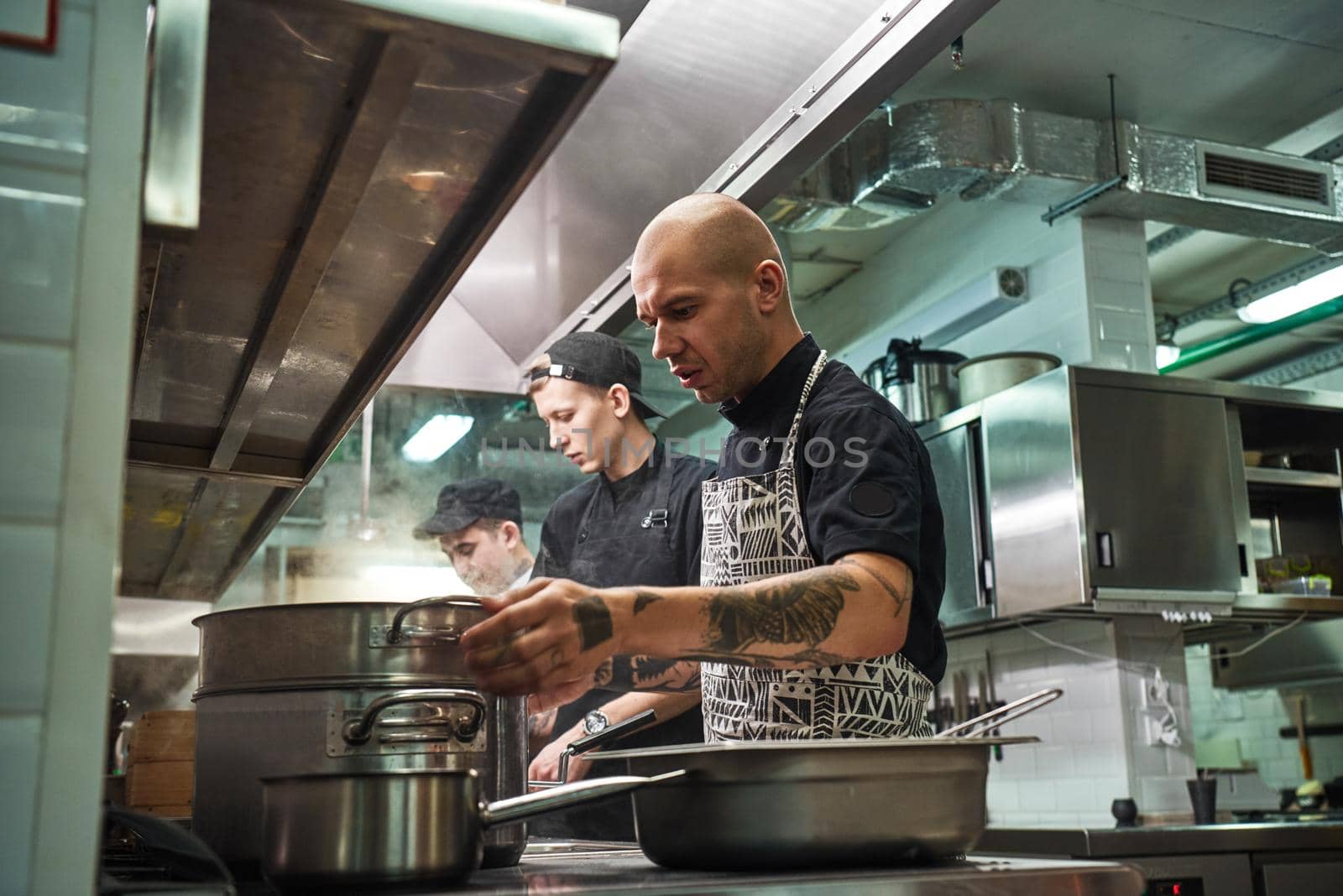 Professional team. Concentrated chef in apron, with tattoos on his arms cooking with his two assistants in a restaurant kitchen by friendsstock
