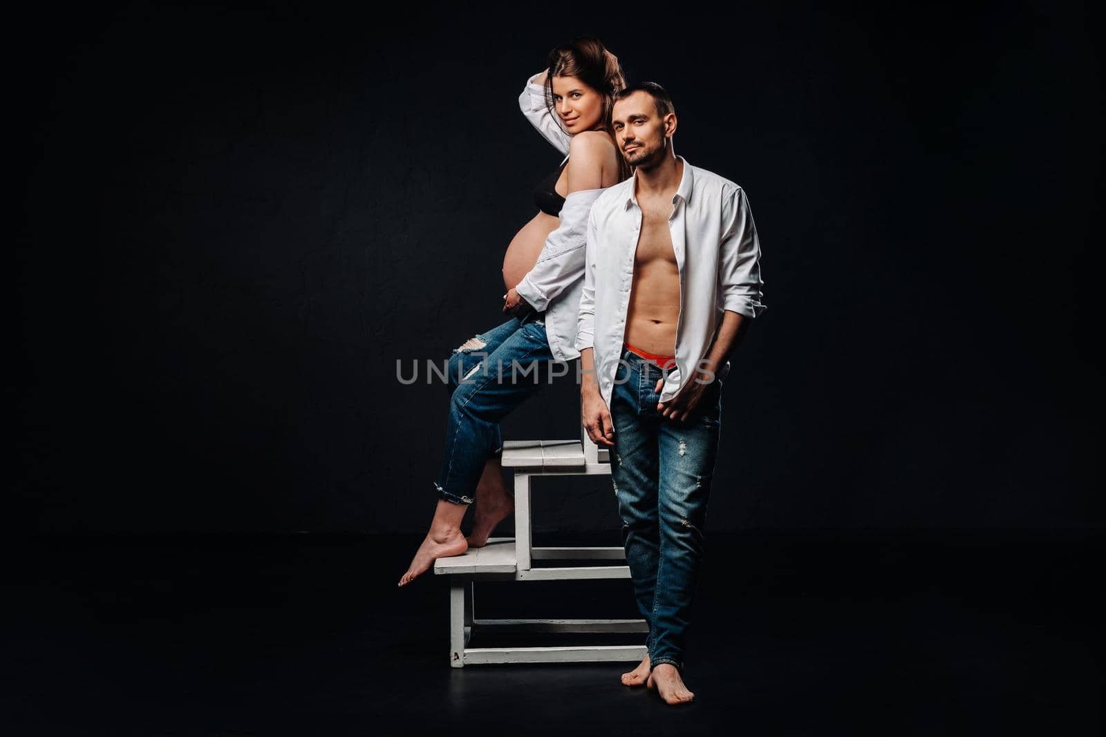 a pregnant woman and a man in a white shirt and jeans in a studio on a black background by Lobachad