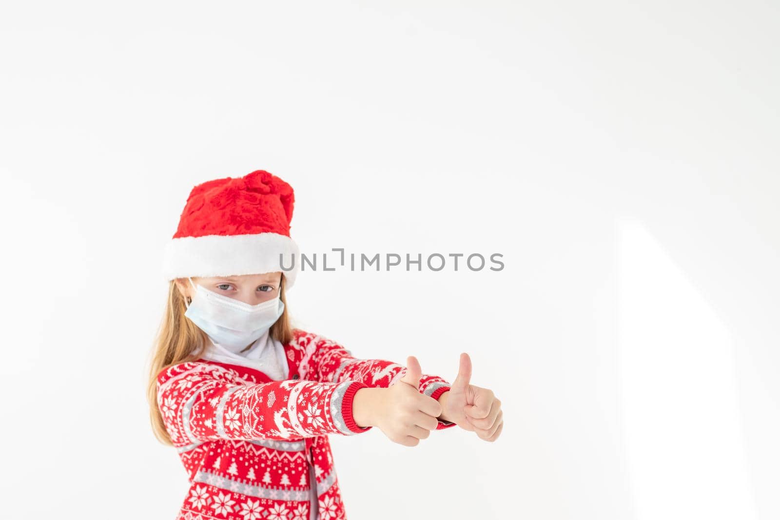 Portrait of a beautiful little girl wearing Santa hat smiling and showing thumbs up, isolated on white background with copy space. celebrating happy New Year.Child safety social distance by YuliaYaspe1979