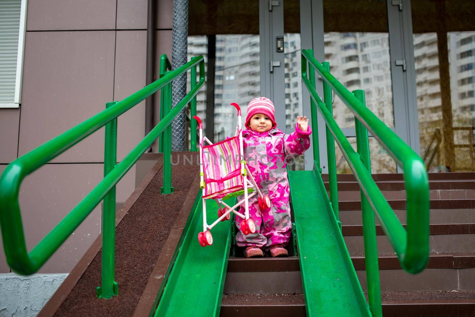 cute toddler with a toy stroller walks along steel railing ramp for wheelchair, carts and strollers. gentle descent from the stairs outdoors