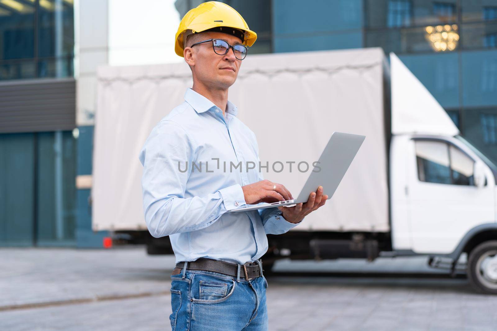 Business. Engineer Worker Protective Helmet Use Laptop Controls Working Process Inspector Supervisor Yellow Hard Hat Glasses Transportation Company Office Building And Truck Background