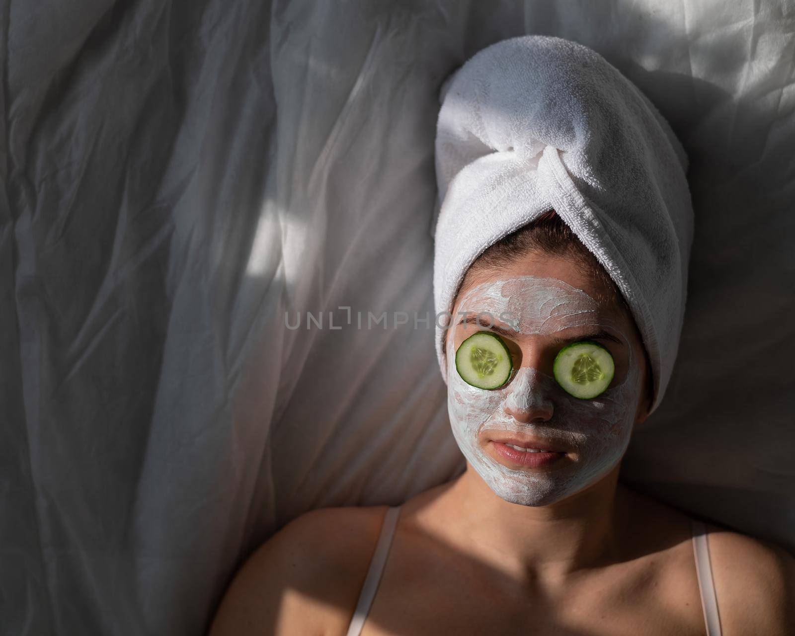A woman with a towel on her hair and in a clay face mask and cucumbers in front of her eyes lies on the sheet by mrwed54