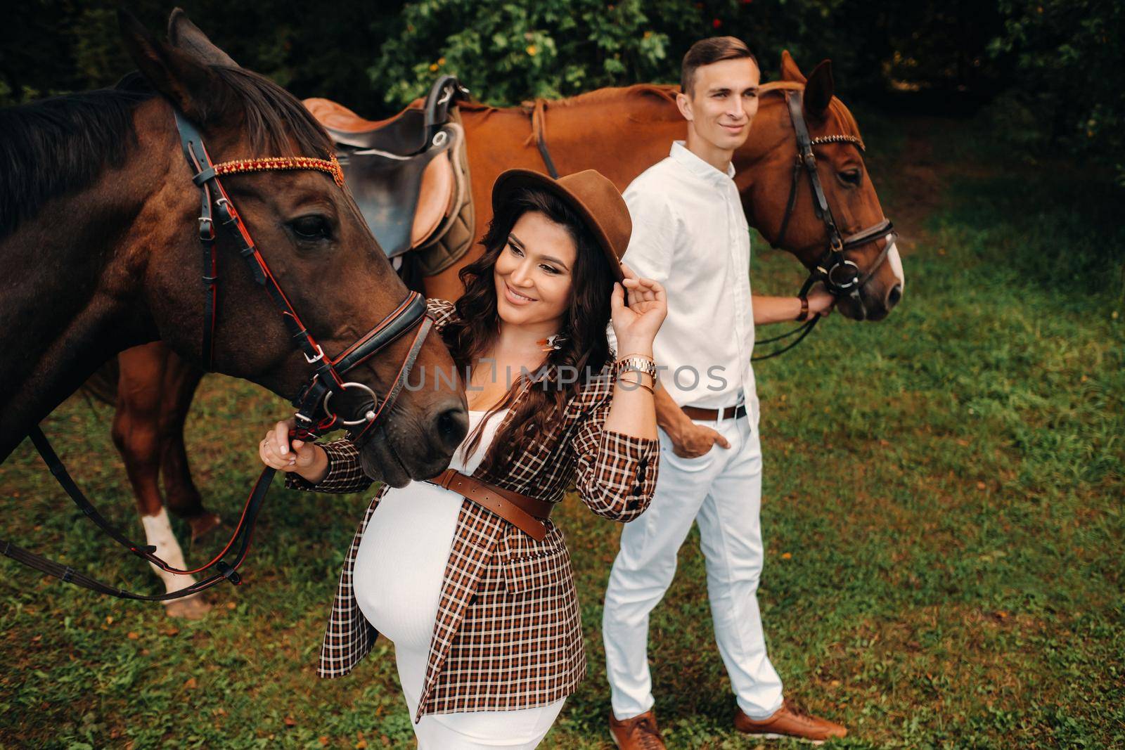 A pregnant woman in a hat with a man in white clothes walking with horses in nature. A family waiting for a child walks in the woods.