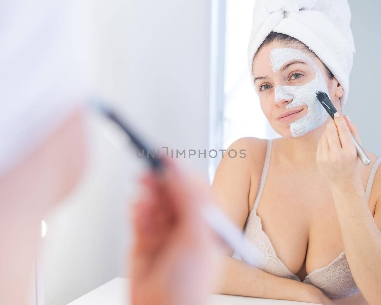 Beautiful caucasian woman with a towel on her hair applies a clay face mask. Taking care of beauty at home by mrwed54