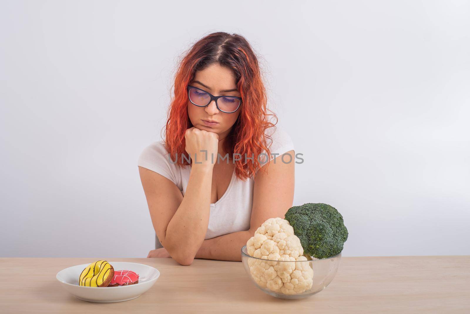 Caucasian woman on a diet dreaming of fast food. Redhead girl chooses between broccoli and donuts on white background