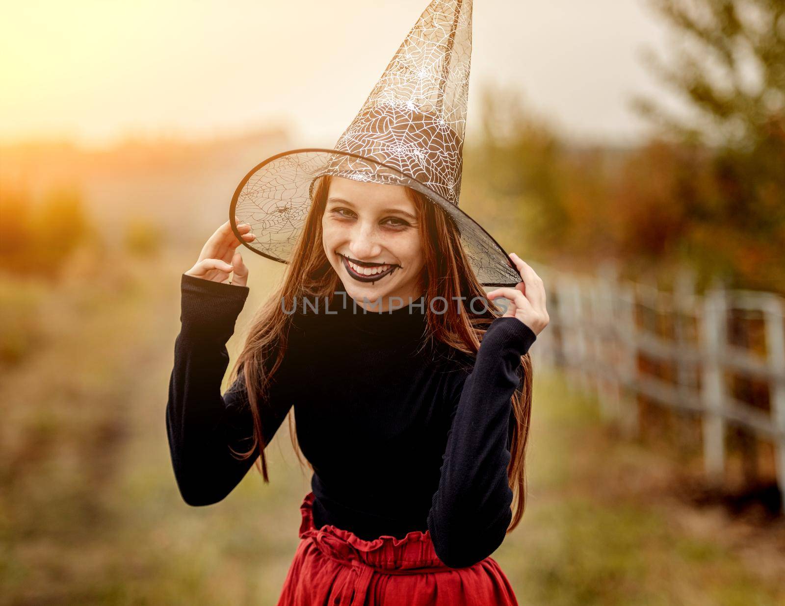 halloween portrait of teenage girl in witch hat smiling at camera on nature background