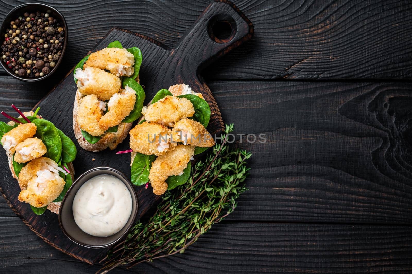 Toasts with Crispy Fried Shrimps Prawns and green salad on a wooden board. Black wooden background. Top view. Copy space.