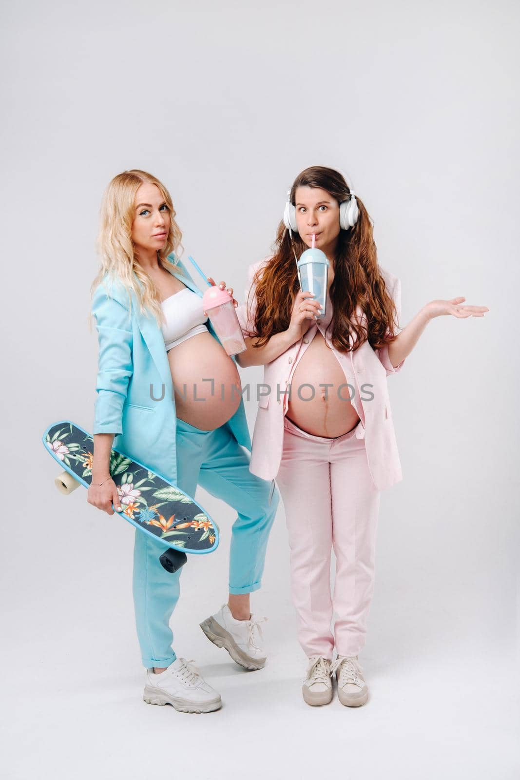 Two pregnant girls in turquoise and pink suits with glasses of juice, a skate and headphones stand on a gray background.