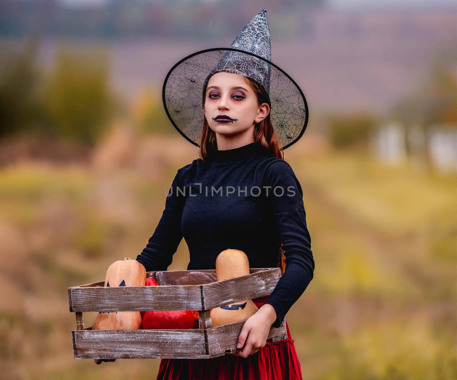 Gothic young girl on halloween in witch costume holding pumpkins box on nature background