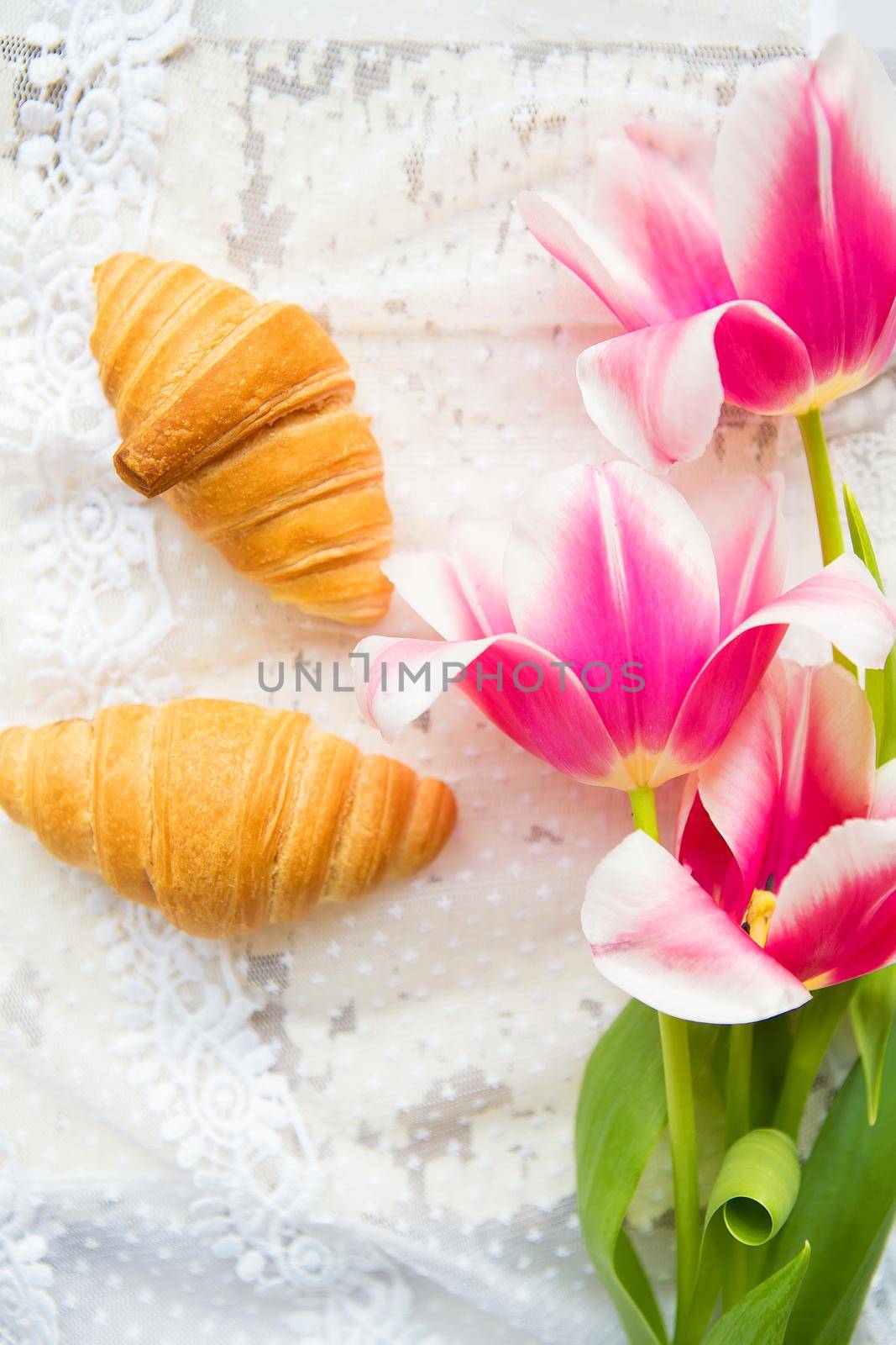 Three croissants and bright pink tulips on lace tablecloth, close-up.