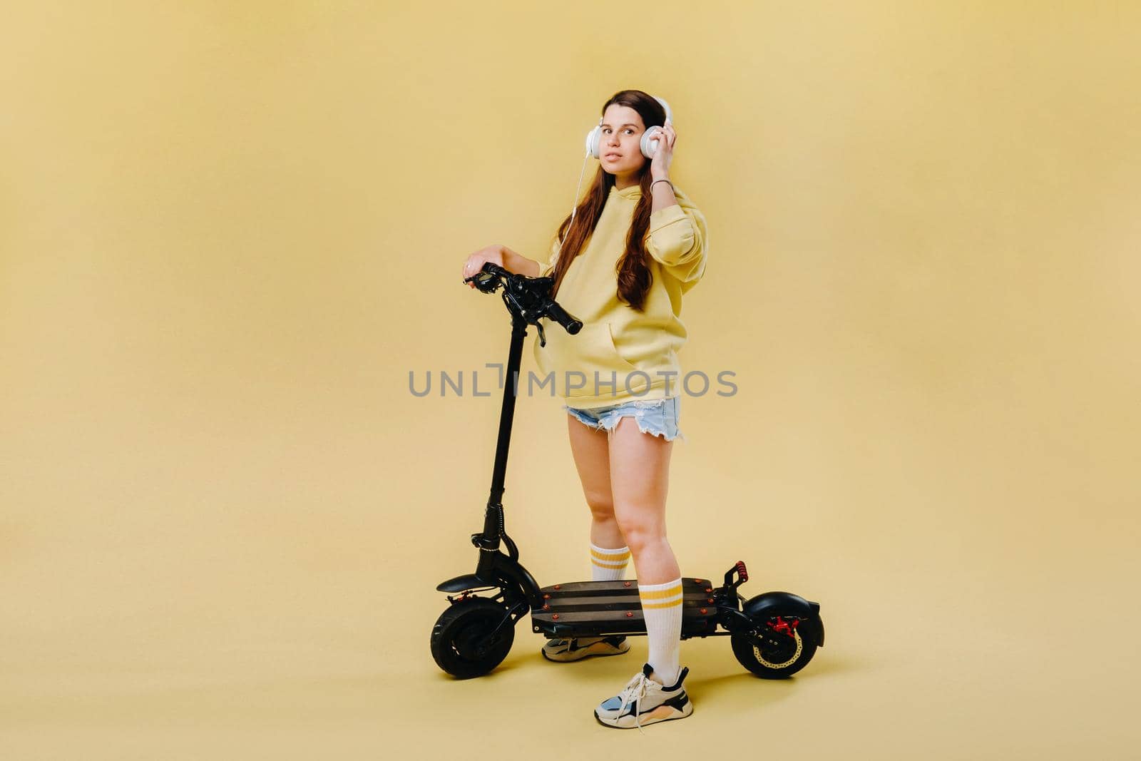 Pregnant girl in yellow clothes and headphones on an electric scooter on an isolated yellow background.