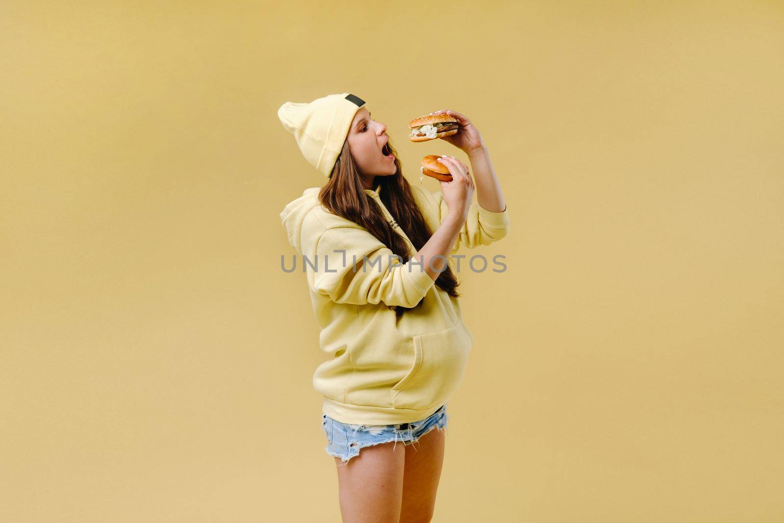 pregnant girl in yellow clothes with hamburgers in her hands on a yellow background.