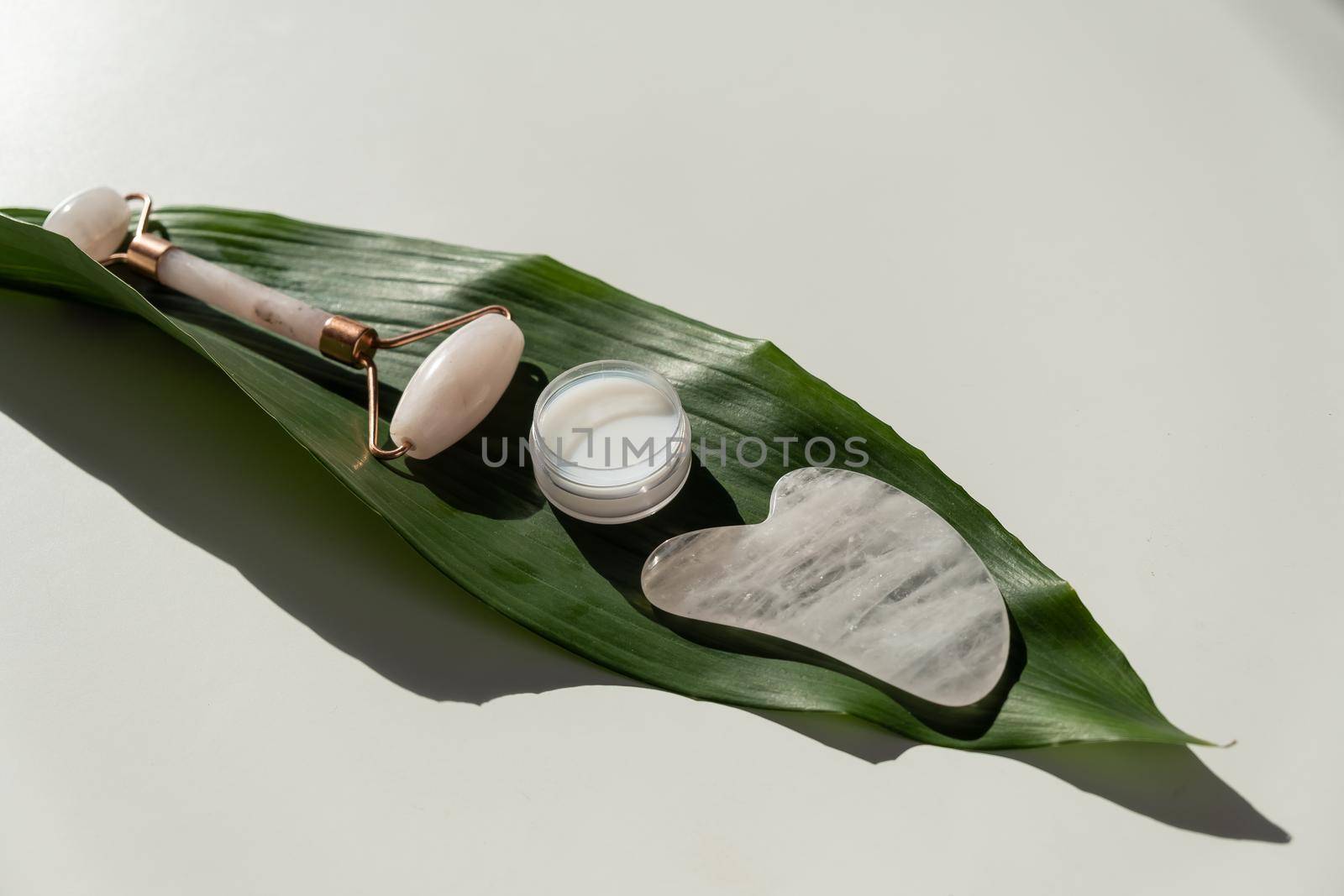 Pink gouache scraper, roller, jar of green leaf cream on a white table. Equipment for self-massage and skin care for the face and neck.