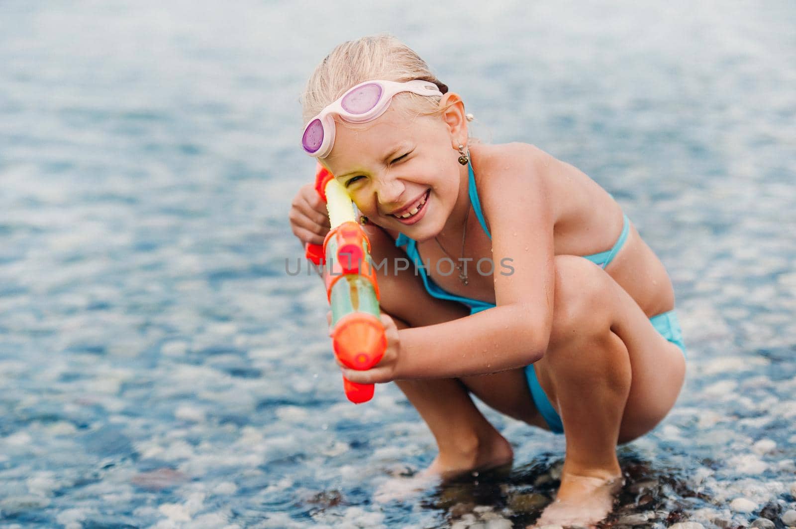 A charming little girl is bathing in the sea and playing with a water gun.A little girl sits in the water and pours water with a water gun.Turkey.