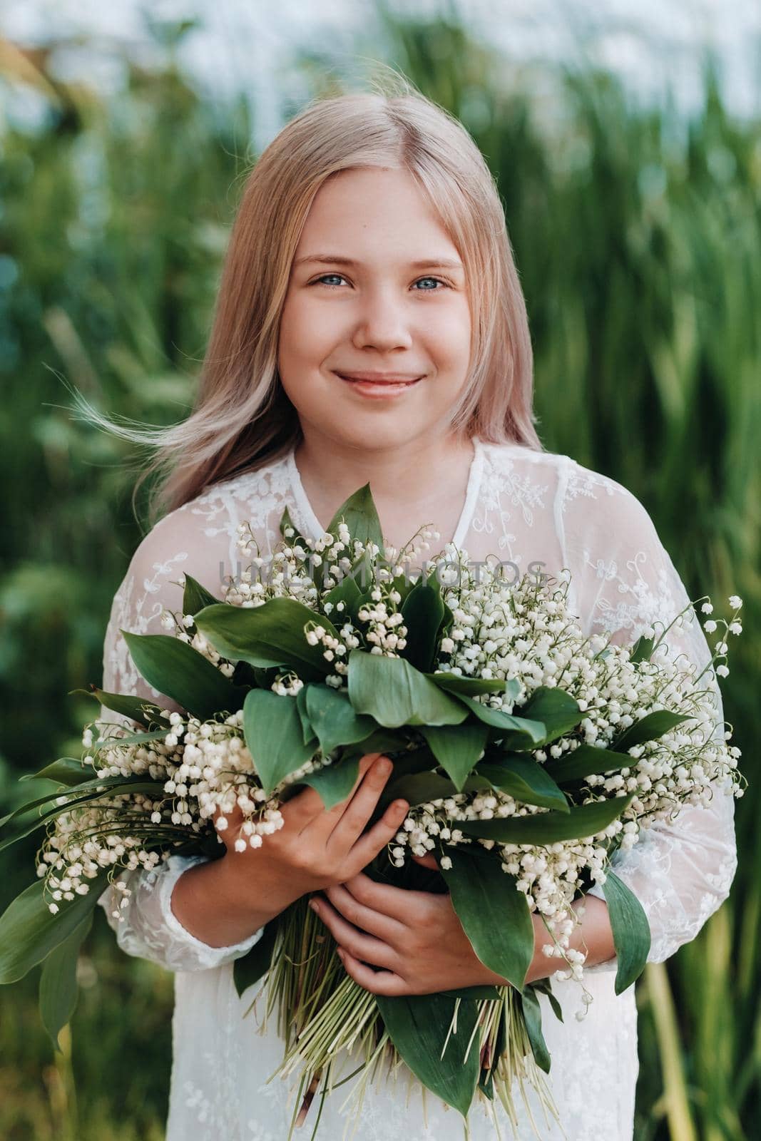 A beautiful nine-year-old blonde girl with long hair in a long white dress, holding a bouquet of lilies of the valley flowers, walking in nature in the Park.Summer, sunset. by Lobachad