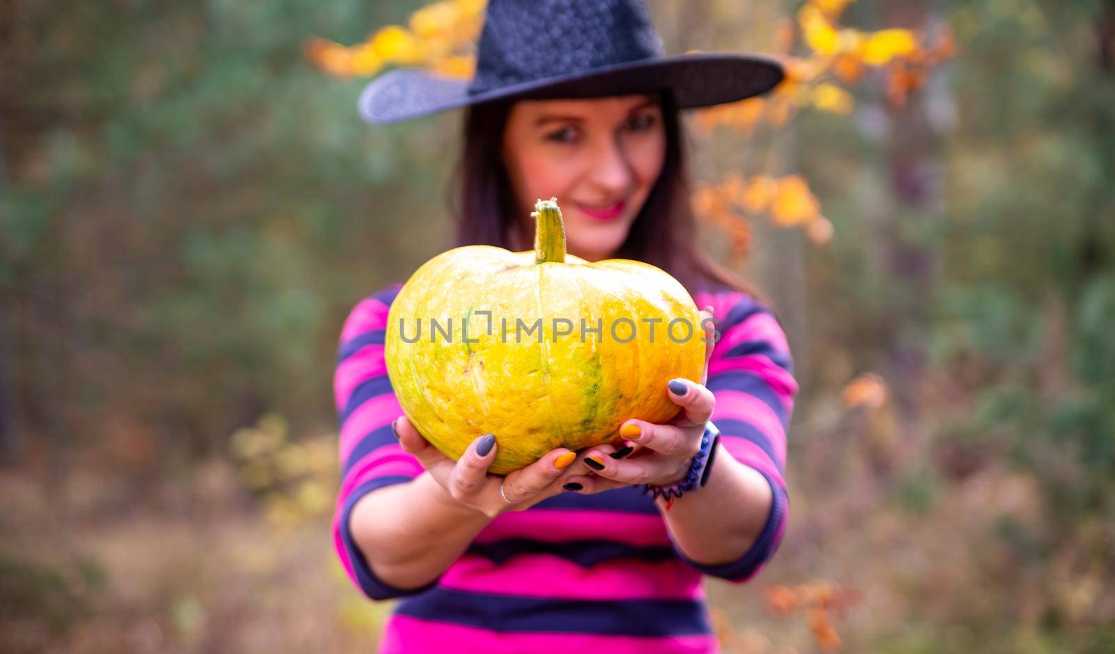 witch holds out a pumpkin to the camera in the autumn forest by Mariaprovector