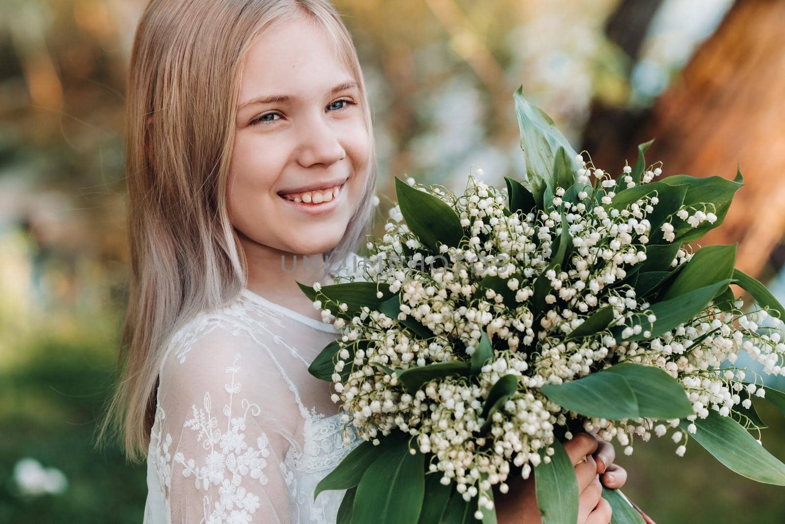 A beautiful nine-year-old blonde girl with long hair in a long white dress, holding a bouquet of lilies of the valley flowers, walking in nature in the Park.Summer, sunset