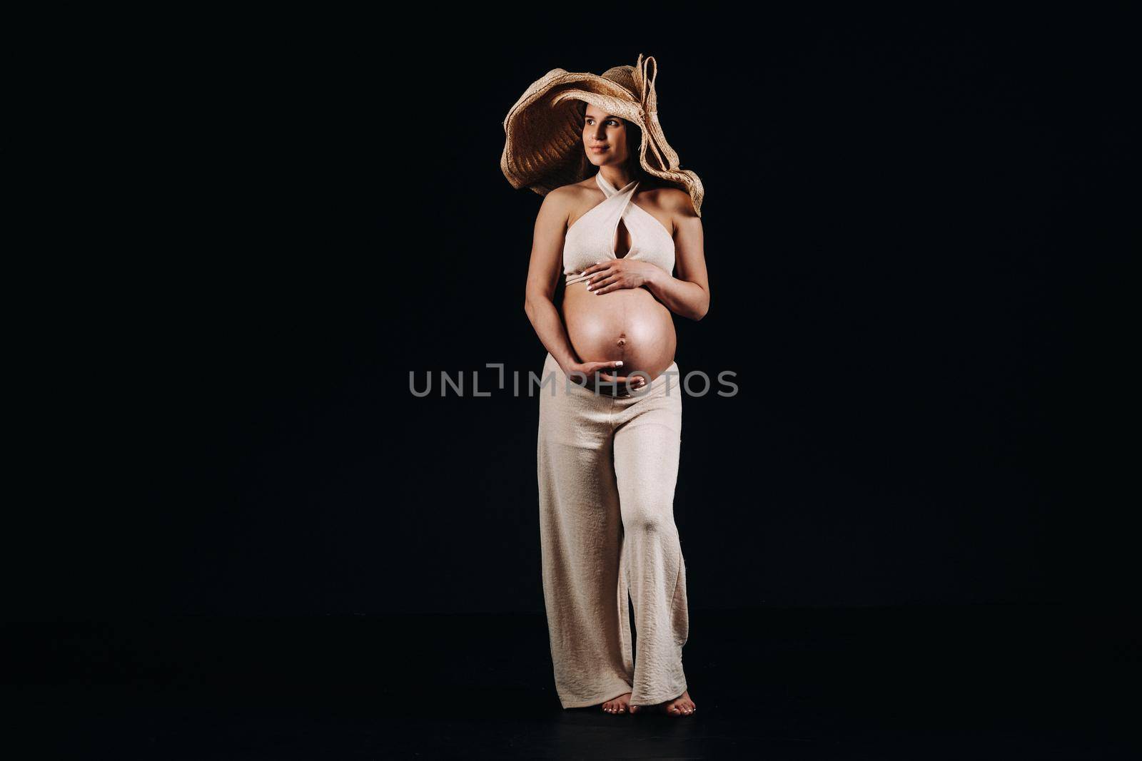 pregnant woman in a straw hat in beige clothes in the studio on a black background.