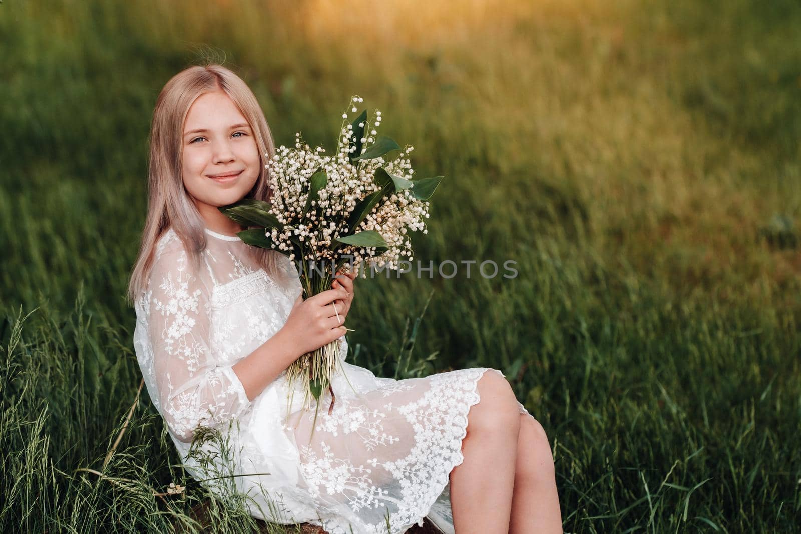 A beautiful nine-year-old blonde girl with long hair in a long white dress, holding a bouquet of lilies of the valley flowers, walking in nature in the Park.Summer, sunset