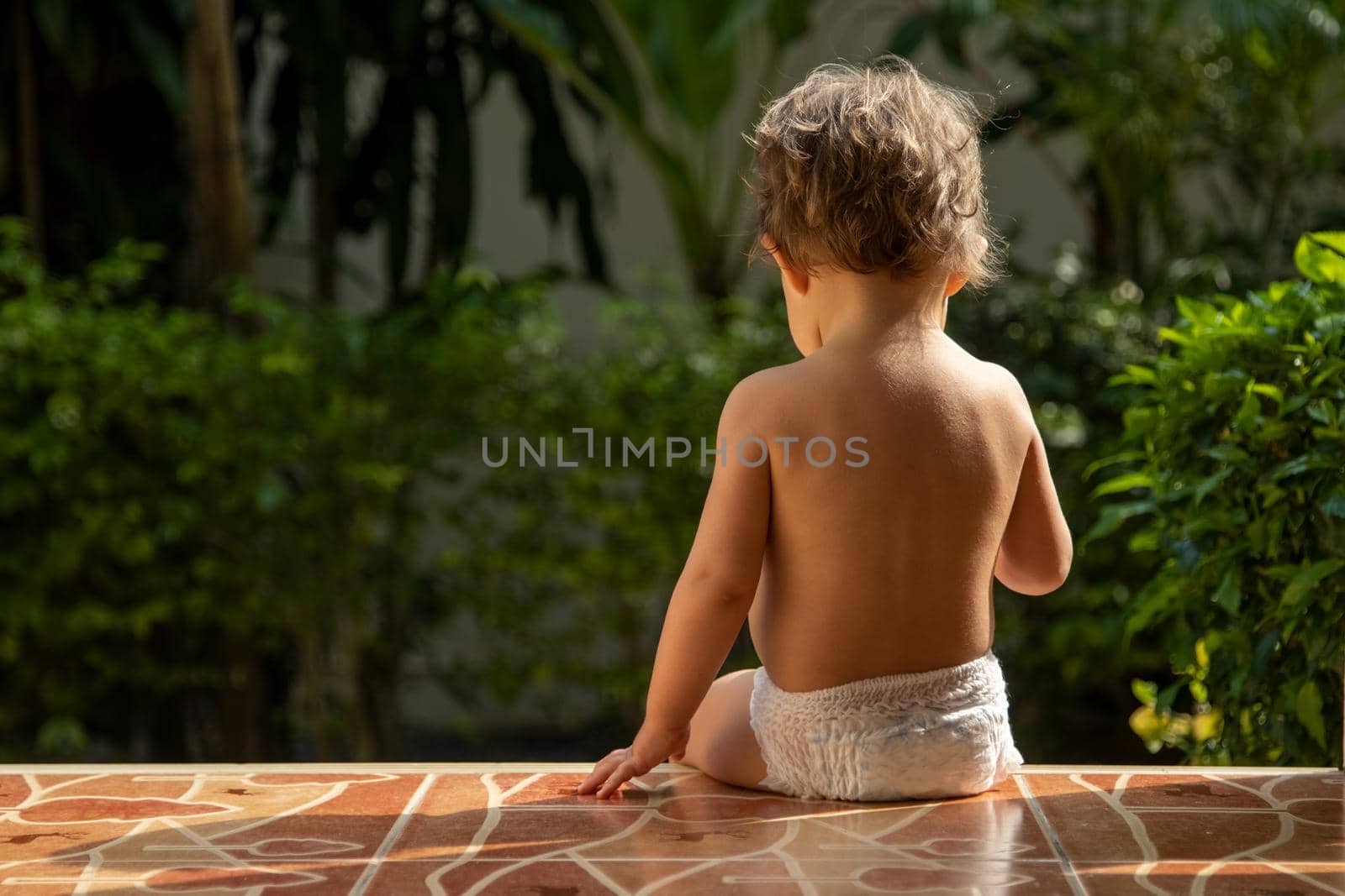 a charming toddler sits on the steps of a house in the sunlight. back view by Mariaprovector