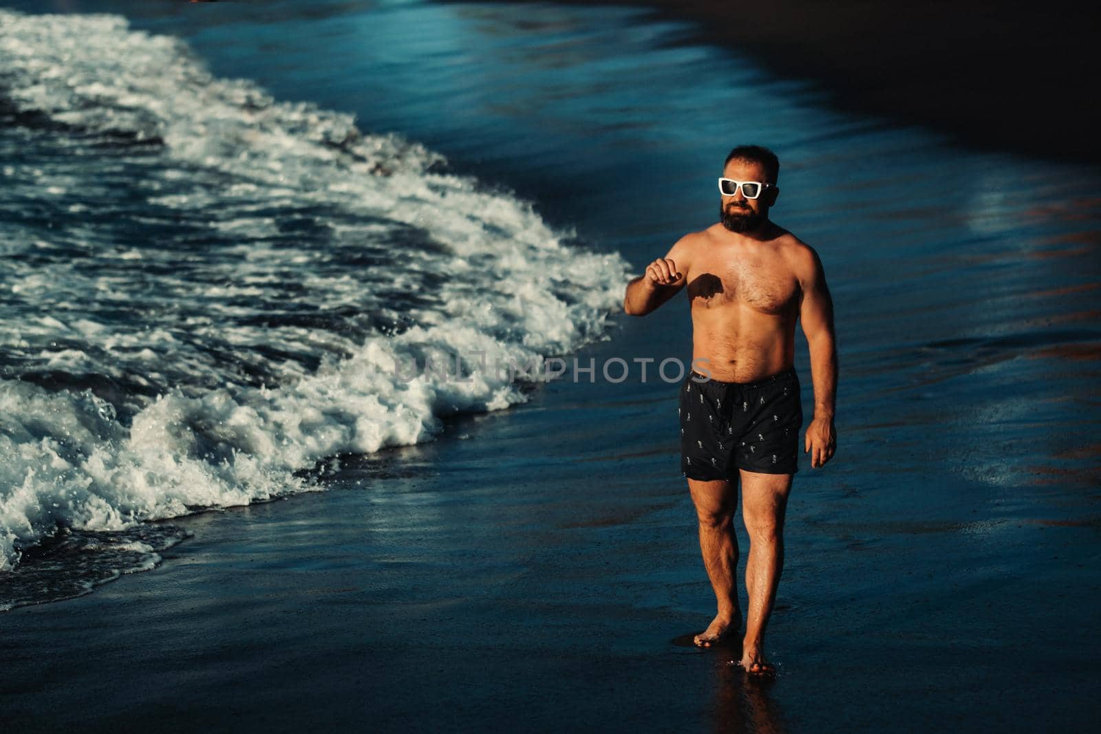 a strong man in black shorts and glasses walks along the beach with black volcanic sand on the island of Tenerife. Spain.