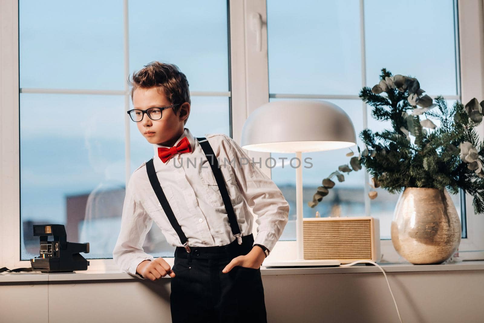 A boy in a shirt with a red bow tie and glasses stands at the window