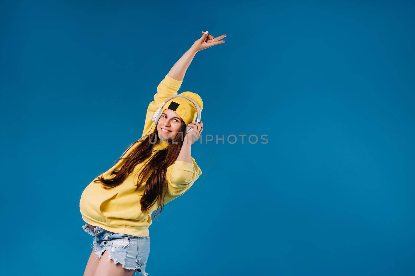 A pregnant girl in a yellow jacket and headphones stands on a blue background.