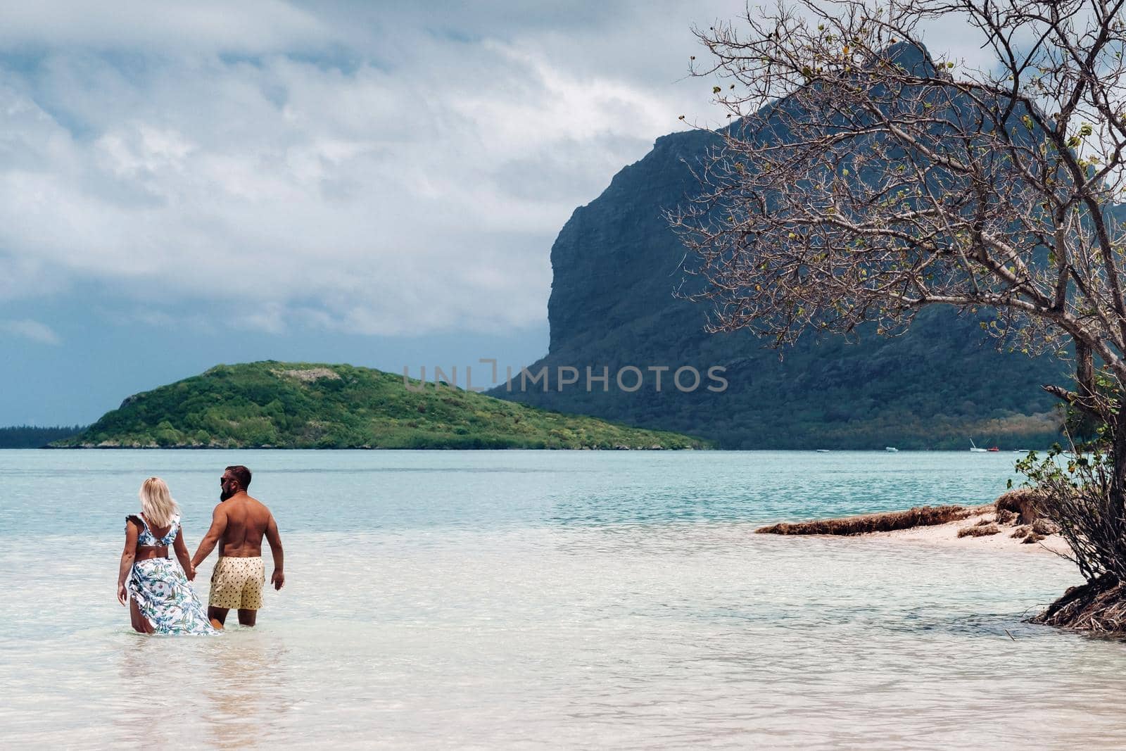 A girl in a swimsuit and a man in shorts stand in the ocean against the backdrop of mount Le Morne on the island of Mauritius.A couple in the water look into the distance of the ocean against the background of mount Le Morne Brabant by Lobachad