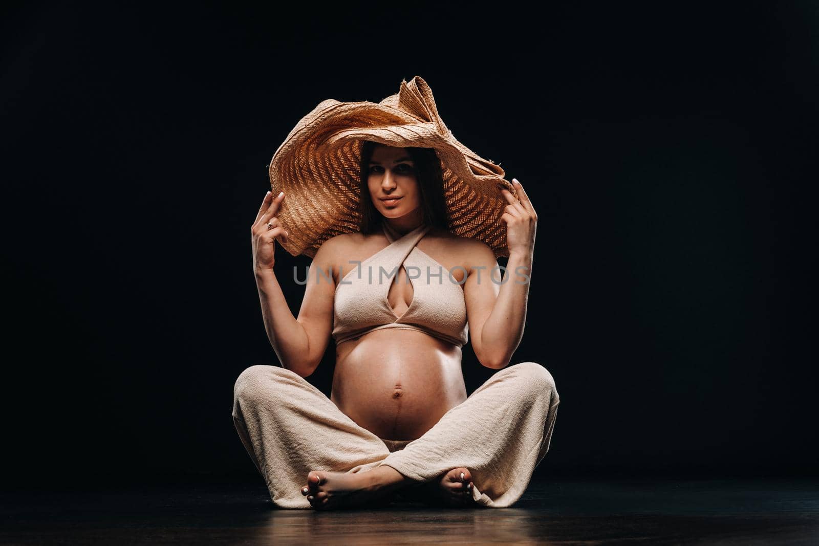 a pregnant woman in a straw hat sits on the floor in beige clothes in a studio on a black background by Lobachad