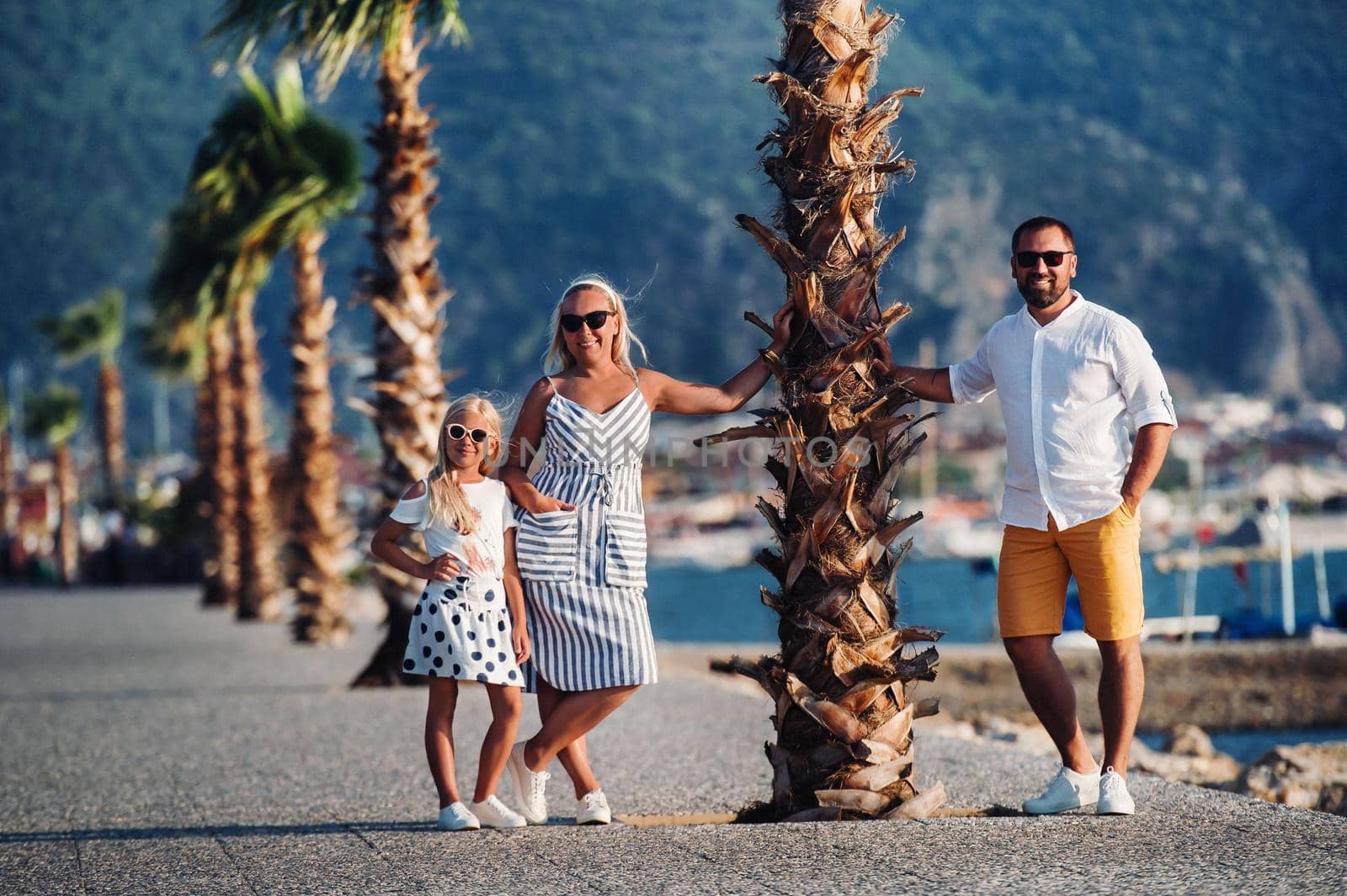 Happy family of mother father and young daughter stands under palm tree with rised hands. Beach holidays concept