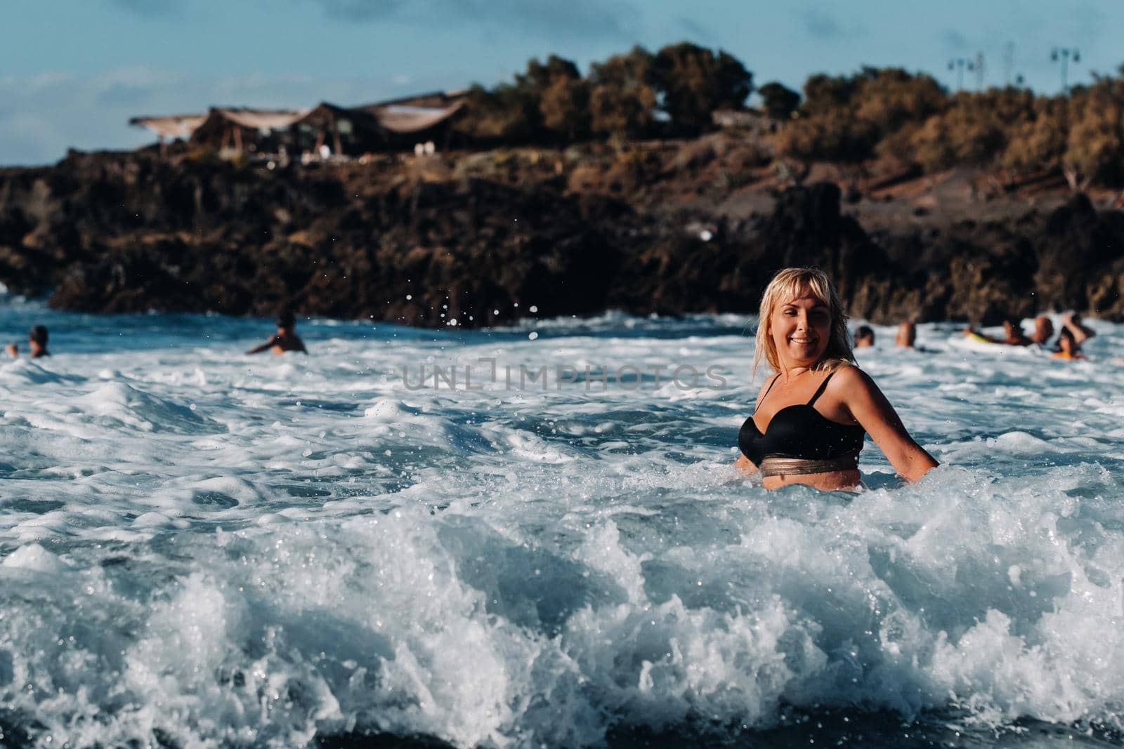 A girl with wet hair and a black swimsuit stands in the white foam of sea water on the island of Tenerife, around a wave with splashes of spray and water drops.Canary Islands.Spain.