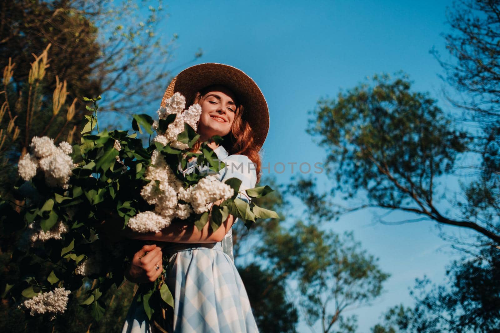 Portrait of a beautiful girl with long hair, a straw hat and a long summer dress with lilac flowers in the garden.