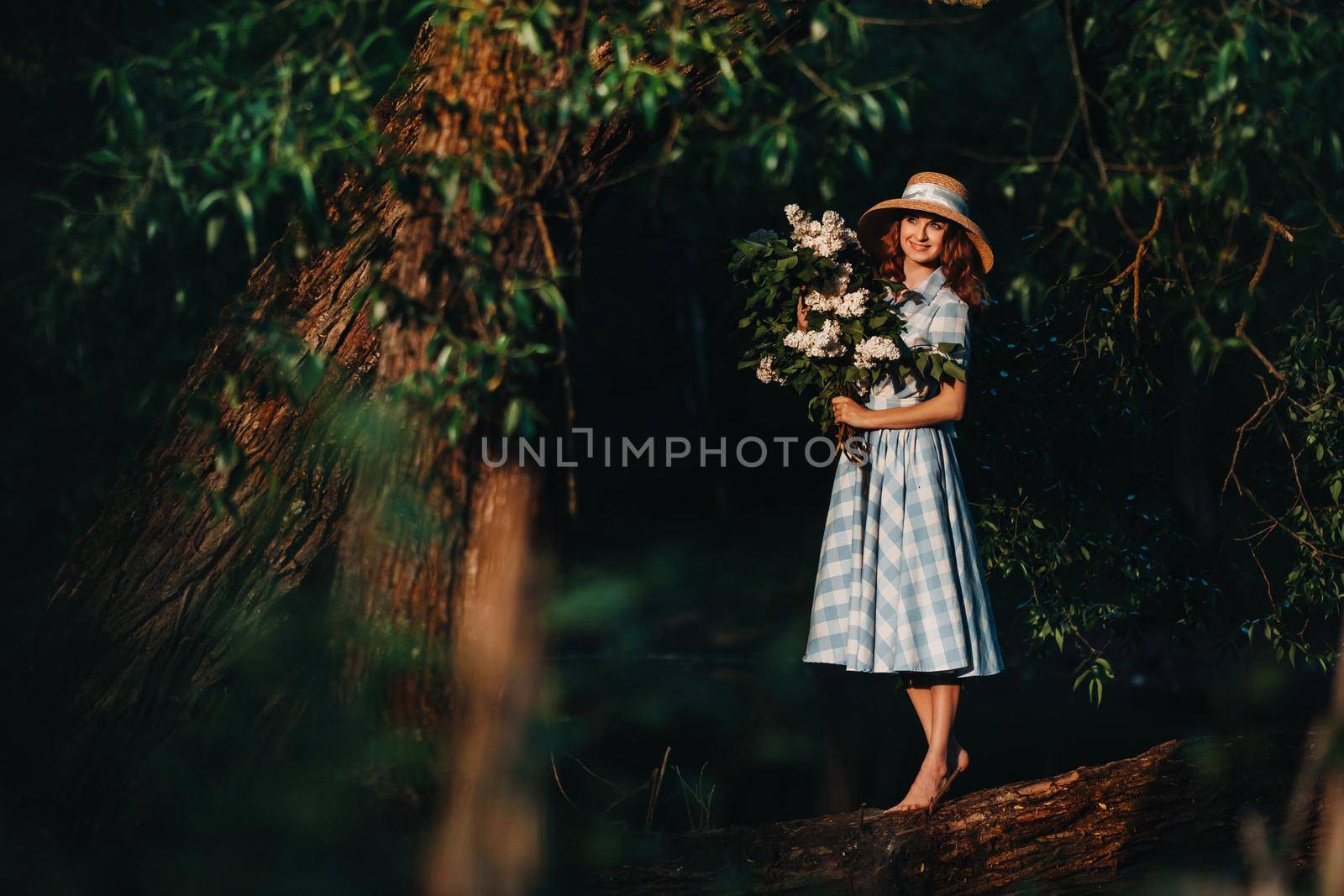 A stylish woman with a straw hat and bare feet poses on a fallen tree with a bouquet of lilacs in a Sunny spring Park. A quiet portrait of a beautiful girl standing in a purple lilac by Lobachad