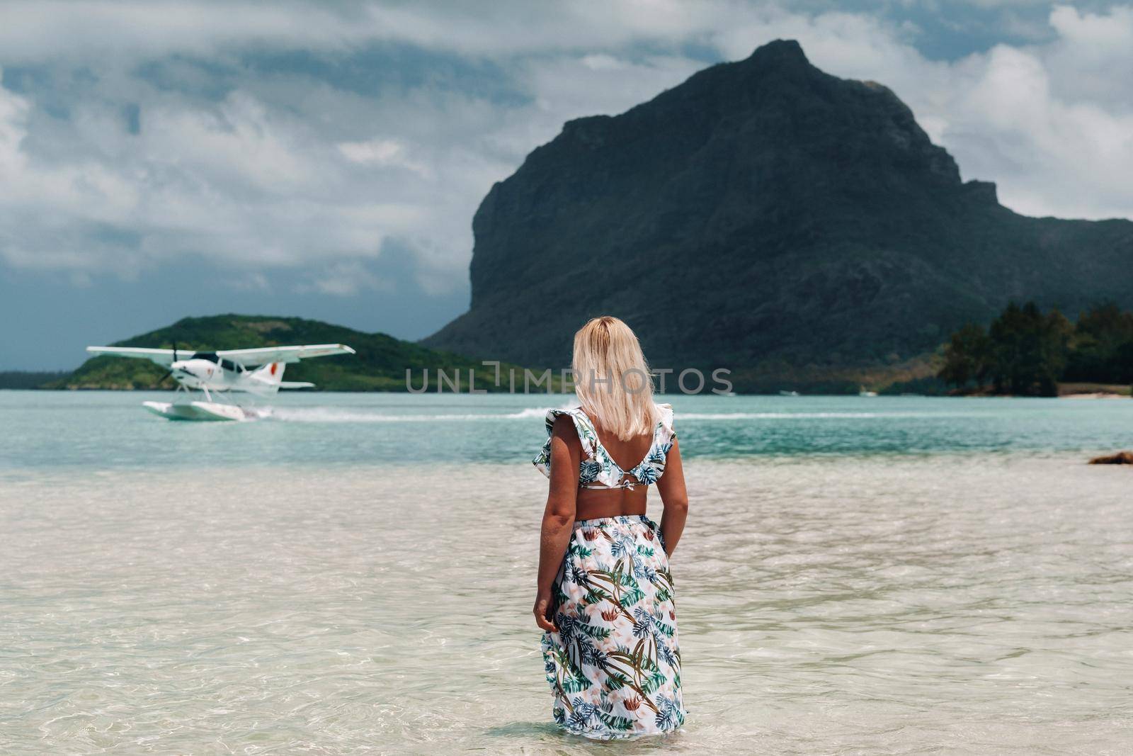 A girl in a swimsuit stands in the ocean and waits for a seaplane against the background of mount Le Morne on the island of Mauritius.A woman in the water looks back at a plane landing on the island of Mauritius.