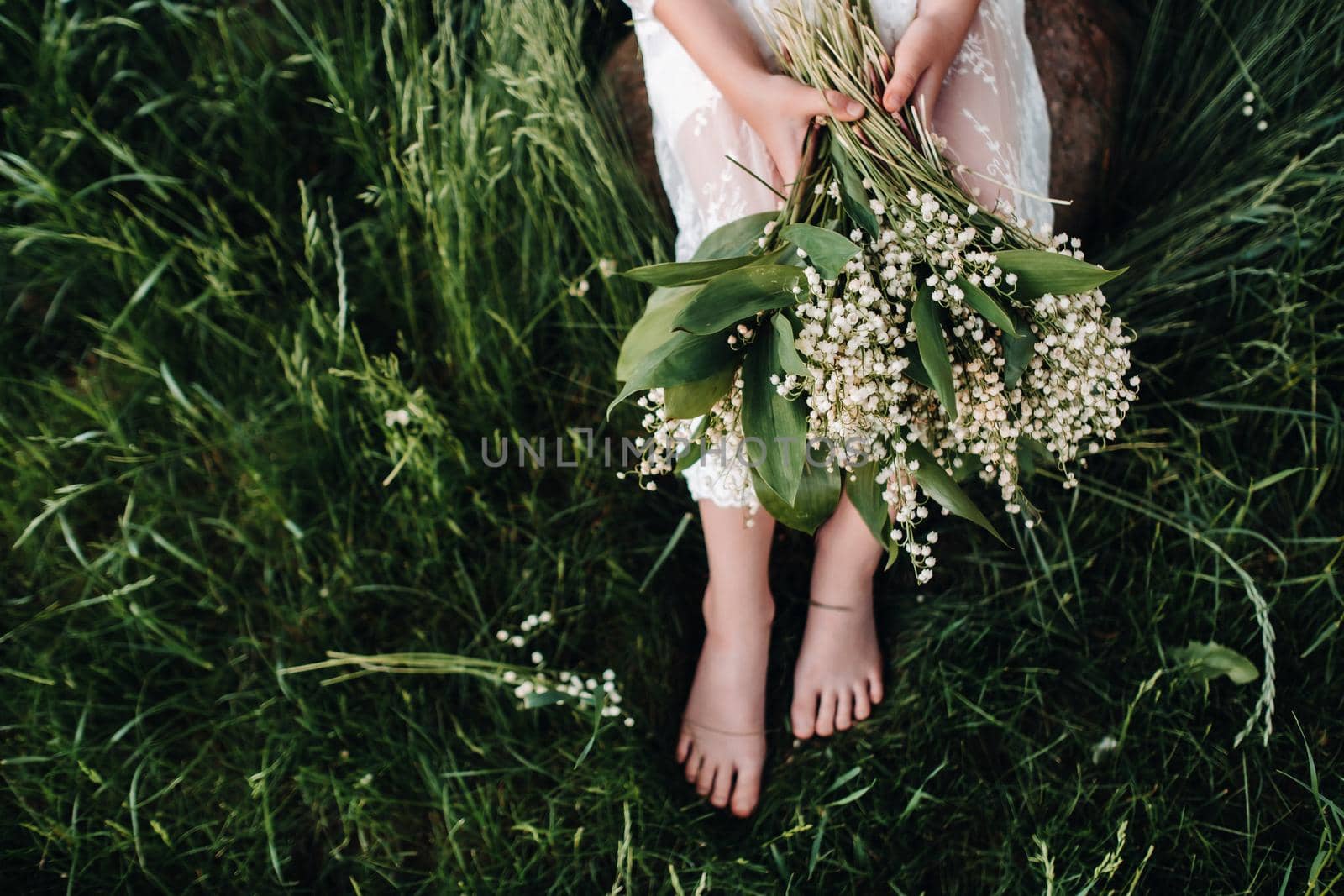 A beautiful nine-year-old blonde in a long white dress, holding a bouquet of lilies of the valley,close-up of a girl holding flowers of the valley.Summer, sunset