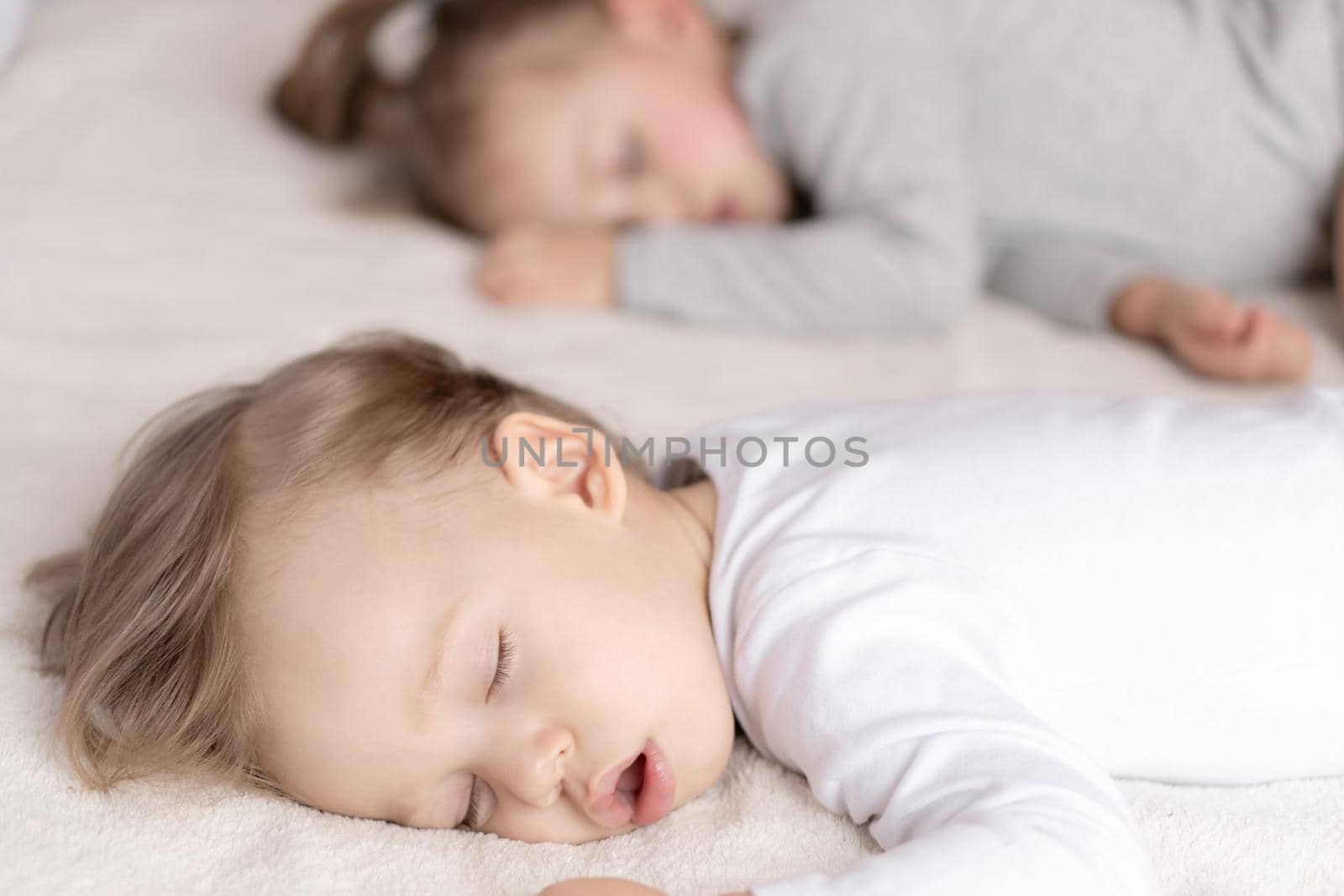 Childhood, sleep, relaxation, family, lifestyle concept - two young children 2 and 3 years old dressed in white and beige bodysuit sleep on a beige and white bed at lunch holding hands top view